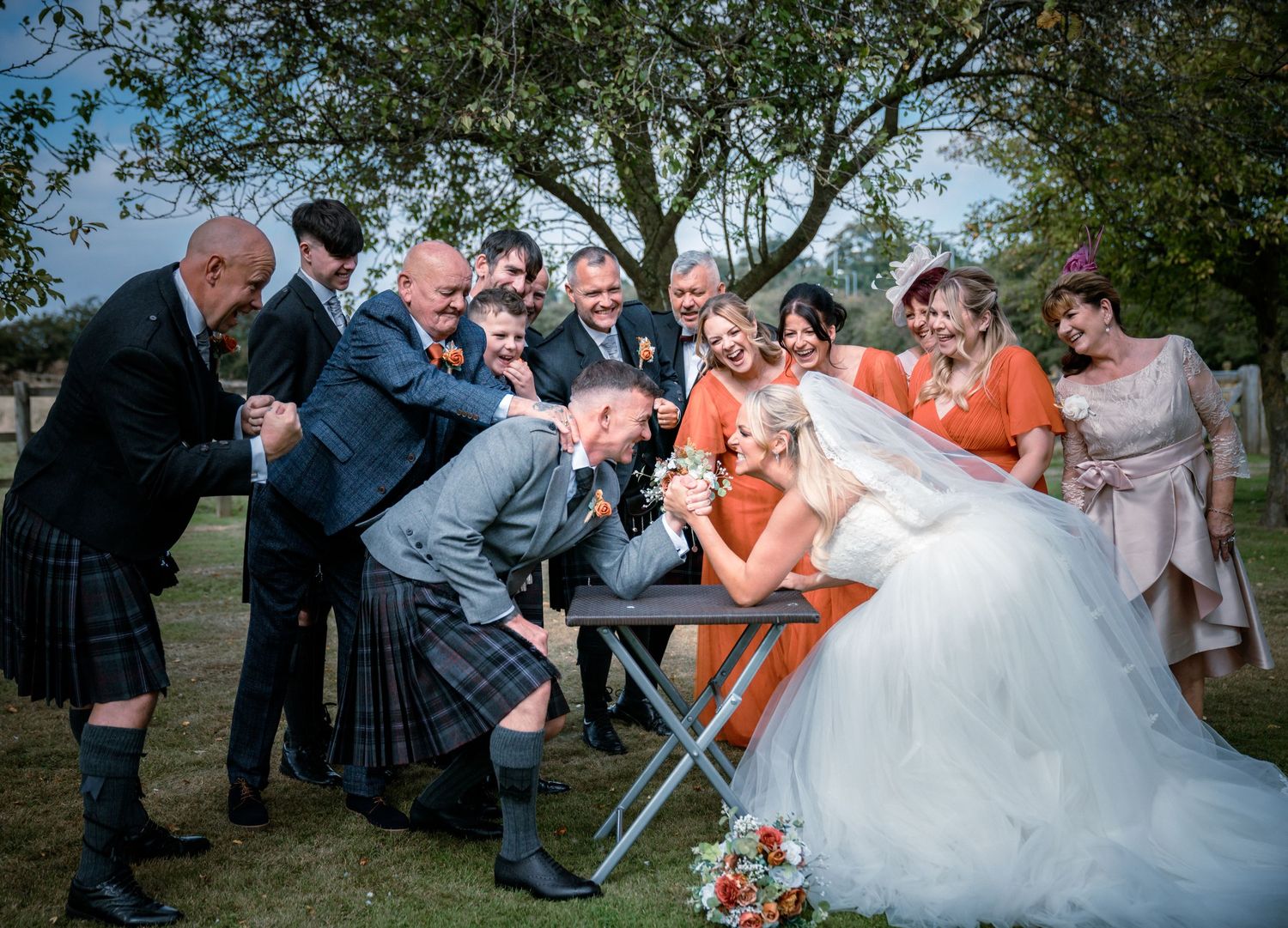 Wedding party cheering as bride and groom arm wrestle at a table outdoors under a large tree.