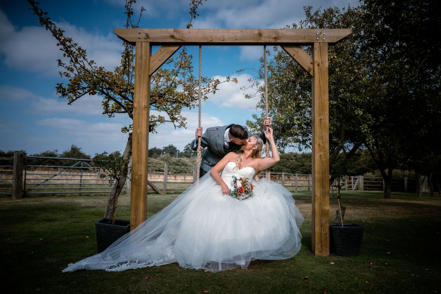 Bride and groom share a romantic kiss on a wooden swing beneath a rustic arch in a garden.