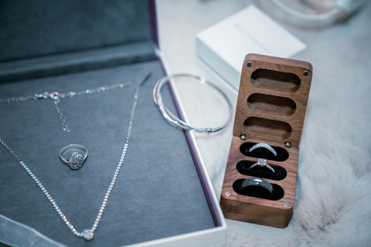 Wedding rings displayed in wooden ring box beside silver necklace and bracelet on soft white fur.