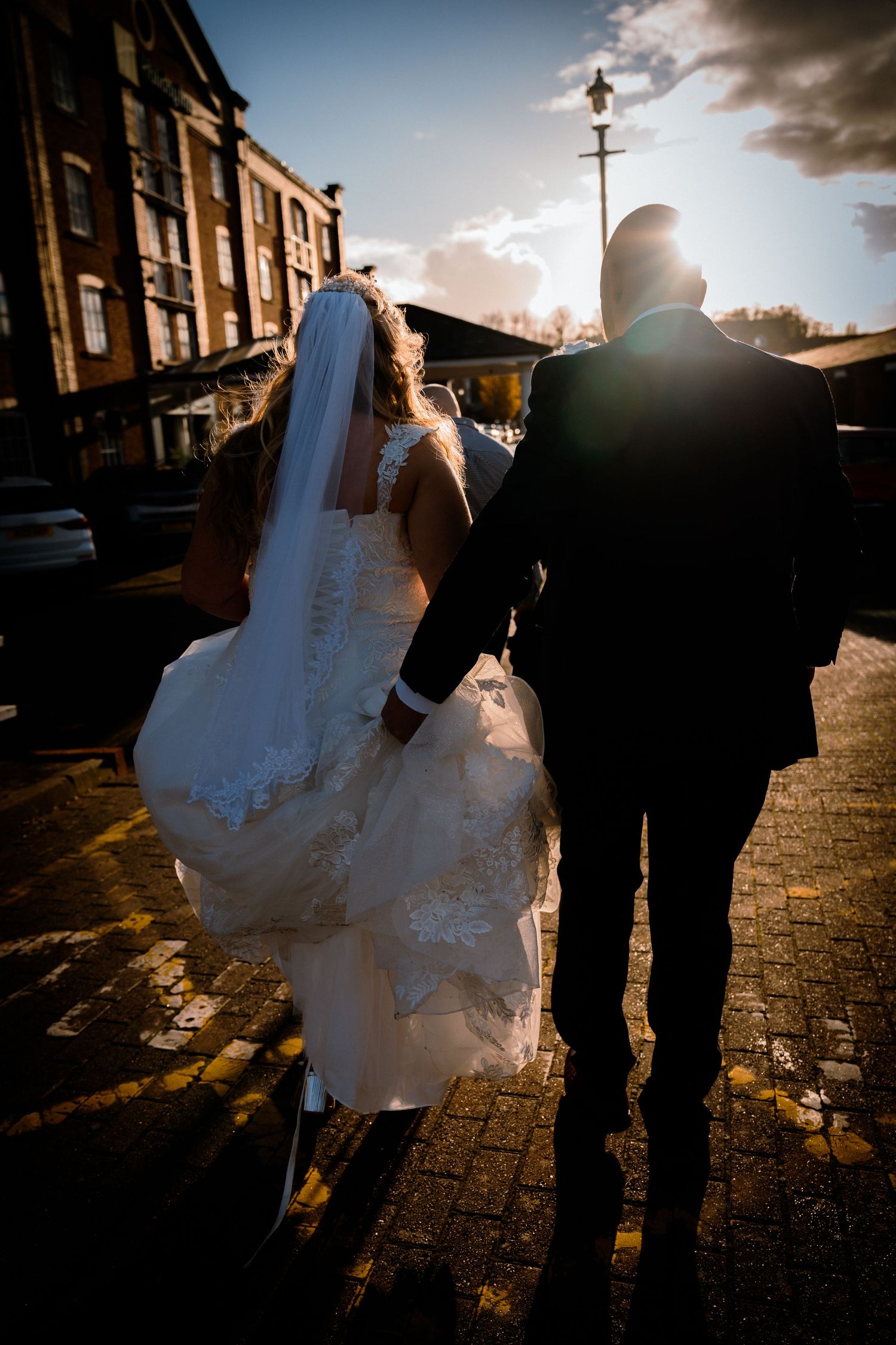 Silhouetted bride and groom walking toward sunset on a cobblestone street with dramatic lens flare.