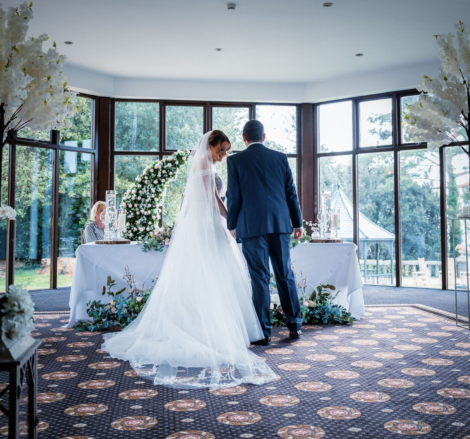 Bride and groom standing at altar during wedding ceremony in bright windowed venue with floral arch.