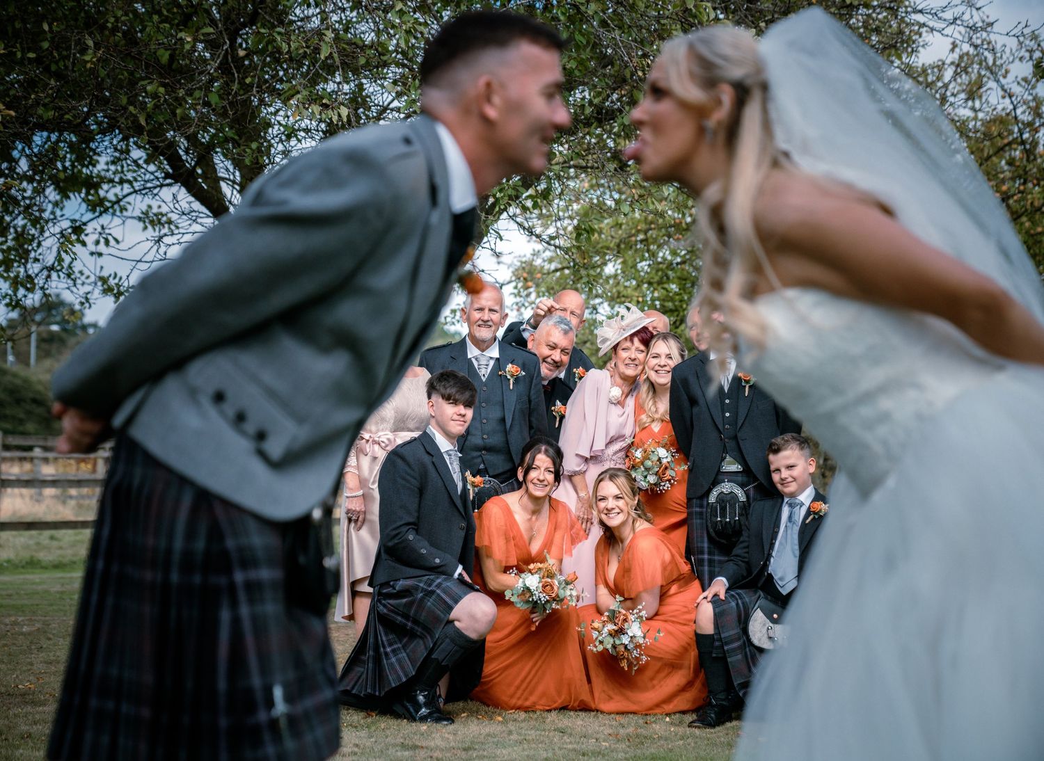 Wedding party posing playfully outdoors with bridesmaids in orange dresses and groom in a kilt.