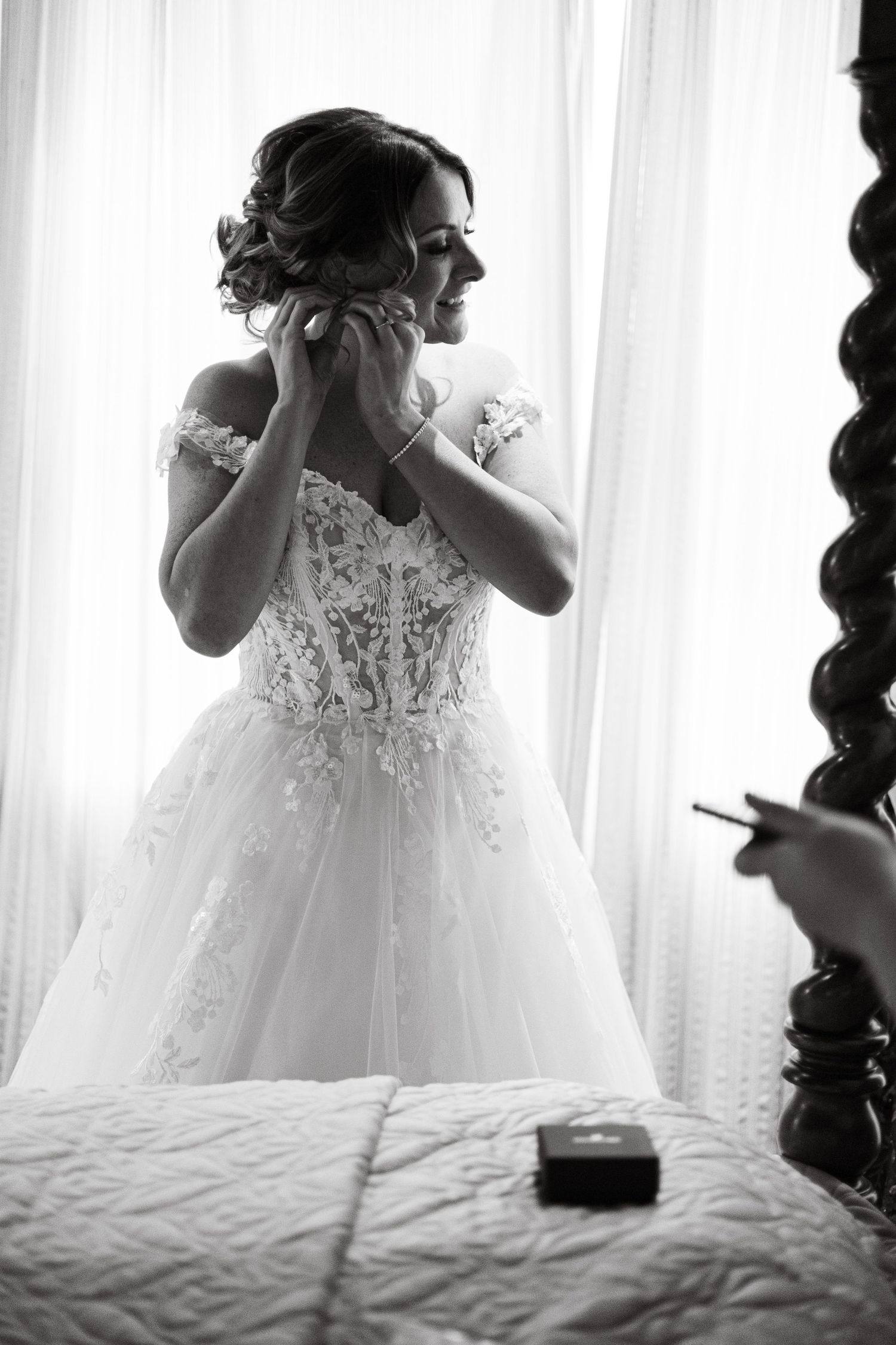 Black and white photo of a bride in a lace ballgown adjusting her earring by a bright window.