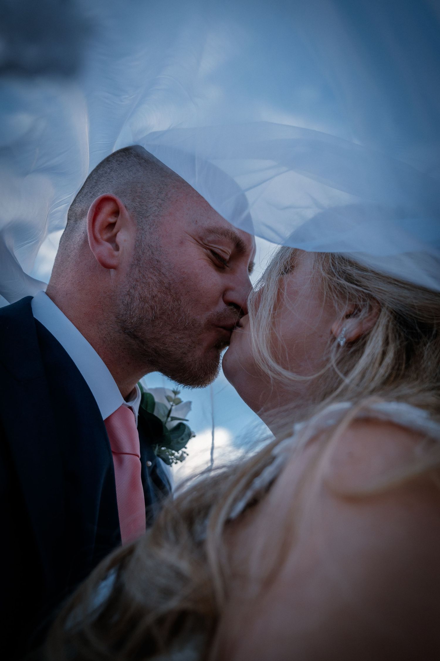 Bride and groom sharing a romantic kiss under a flowing veil against a dramatic cloudy sky.