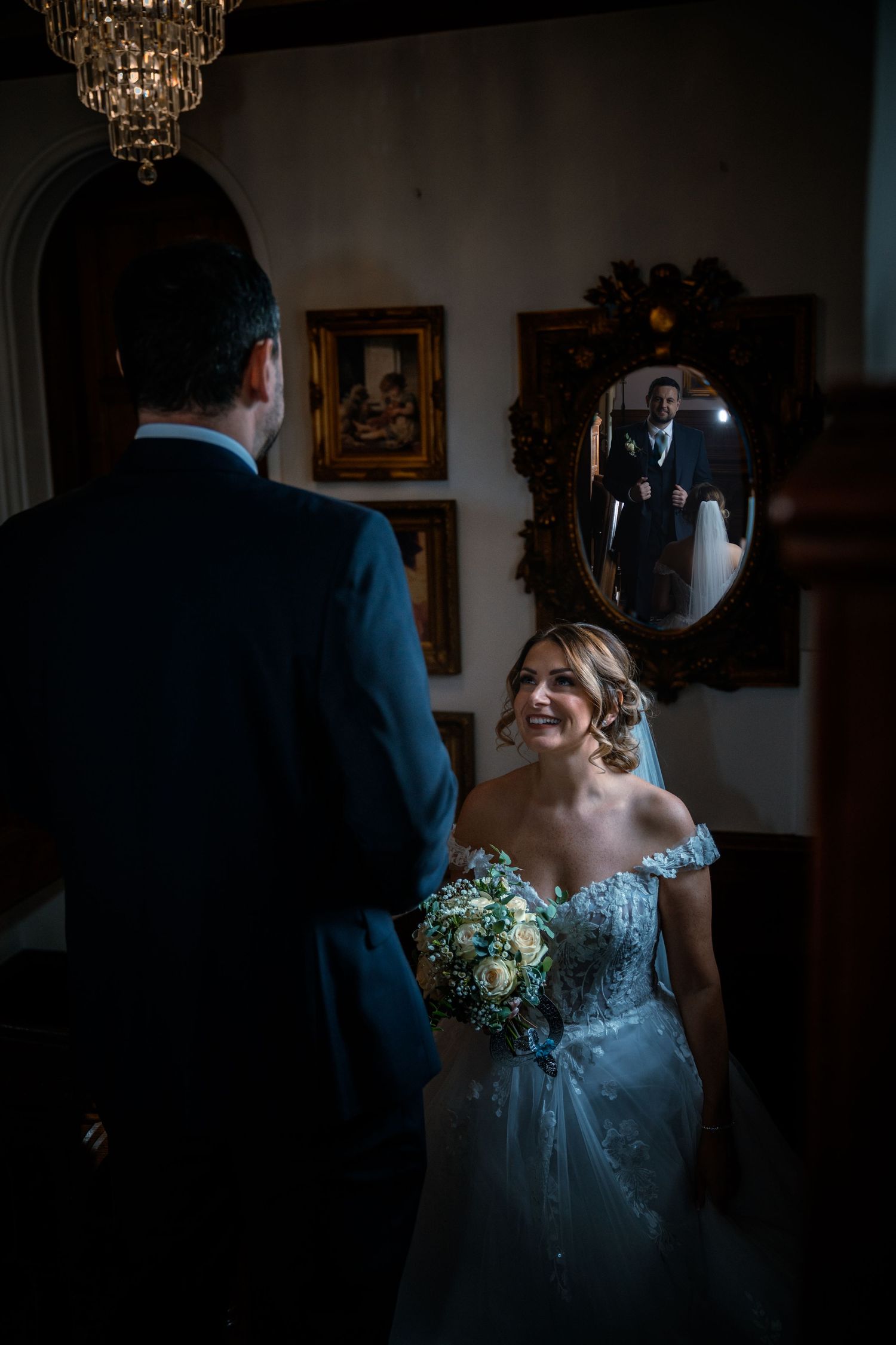Smiling bride in off-shoulder gown holding bouquet faces groom reflected in ornate mirror.