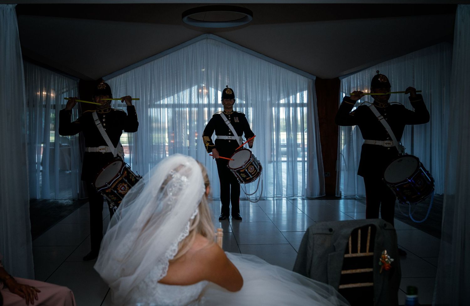 Bride in white gown and veil watches a military band perform trumpets and drums in a grand hall.