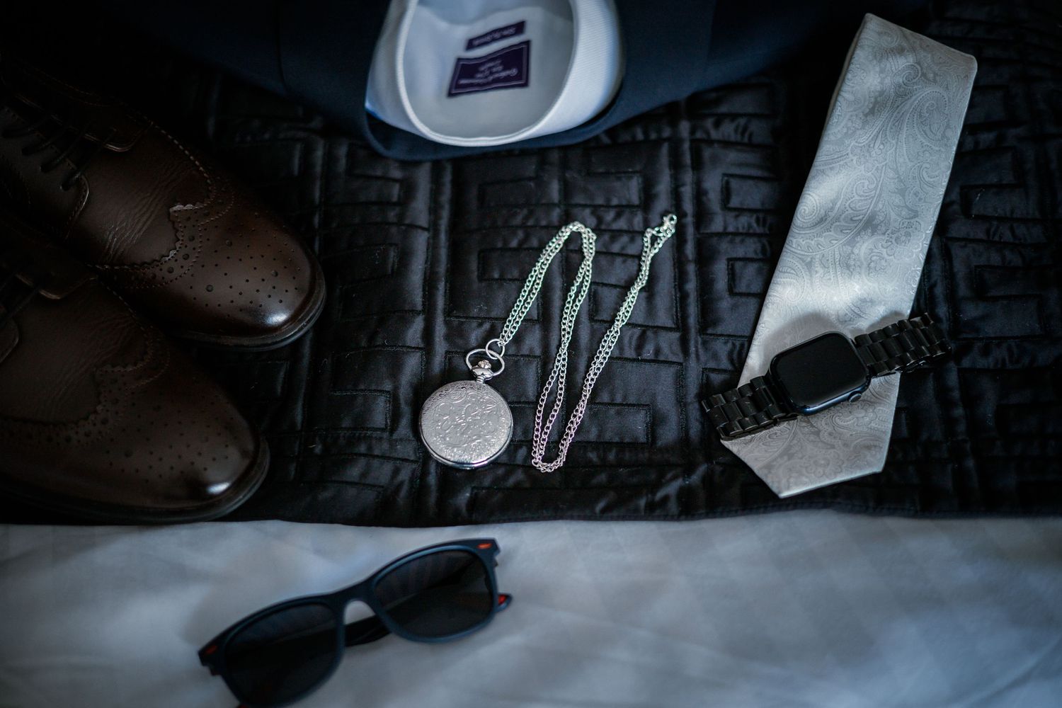 Groom wedding flat lay featuring pocket watch, sunglasses, silver tie, and brown leather shoes.