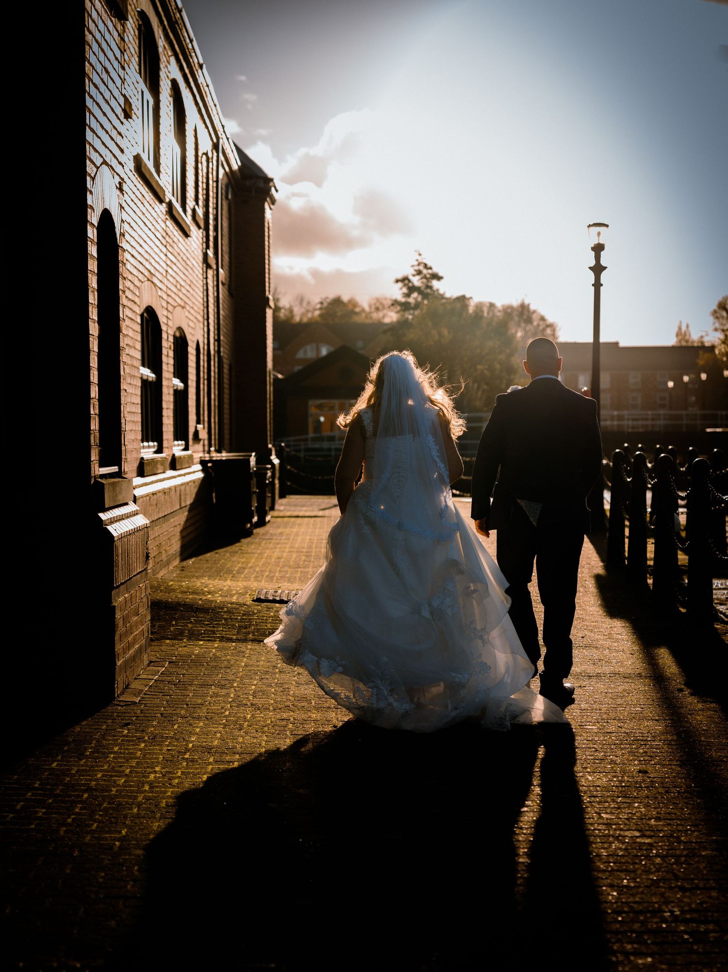 Bride and groom walking in golden sunset light beside a brick building, casting long romantic shadows.