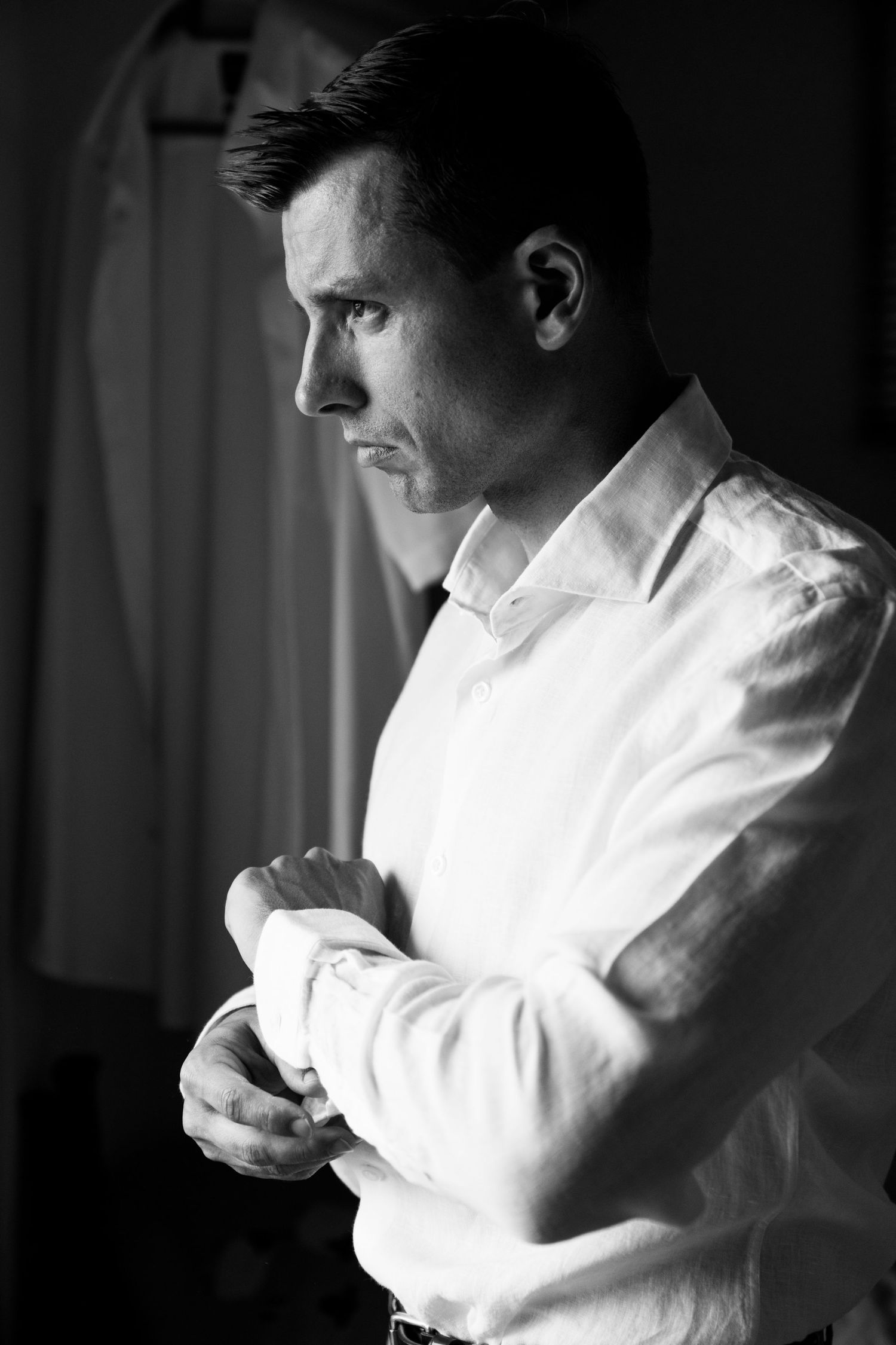 Black and white portrait of a groom buttoning his shirt cuff while gazing thoughtfully out a window.