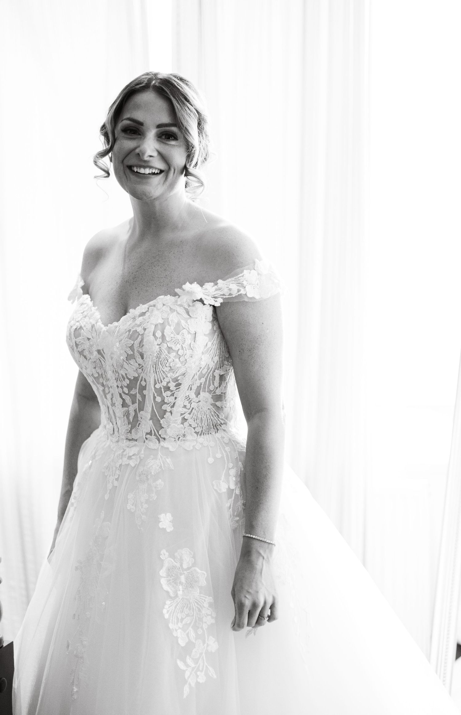 Black and white portrait of smiling bride in off-shoulder lace wedding gown standing by window.