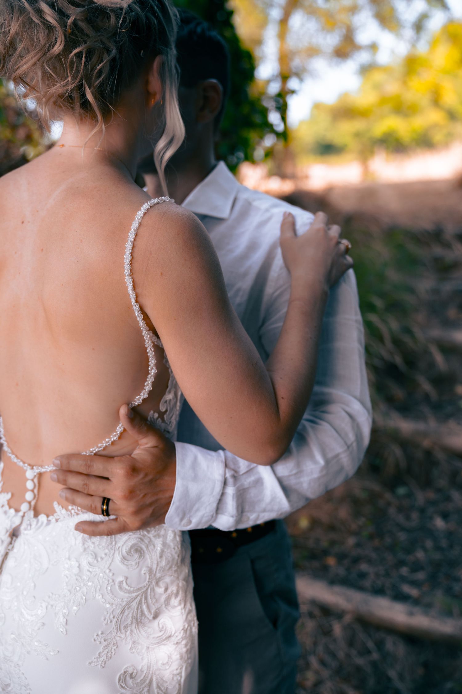 Groom embracing bride from behind showing the open lace back of her wedding dress outdoors.