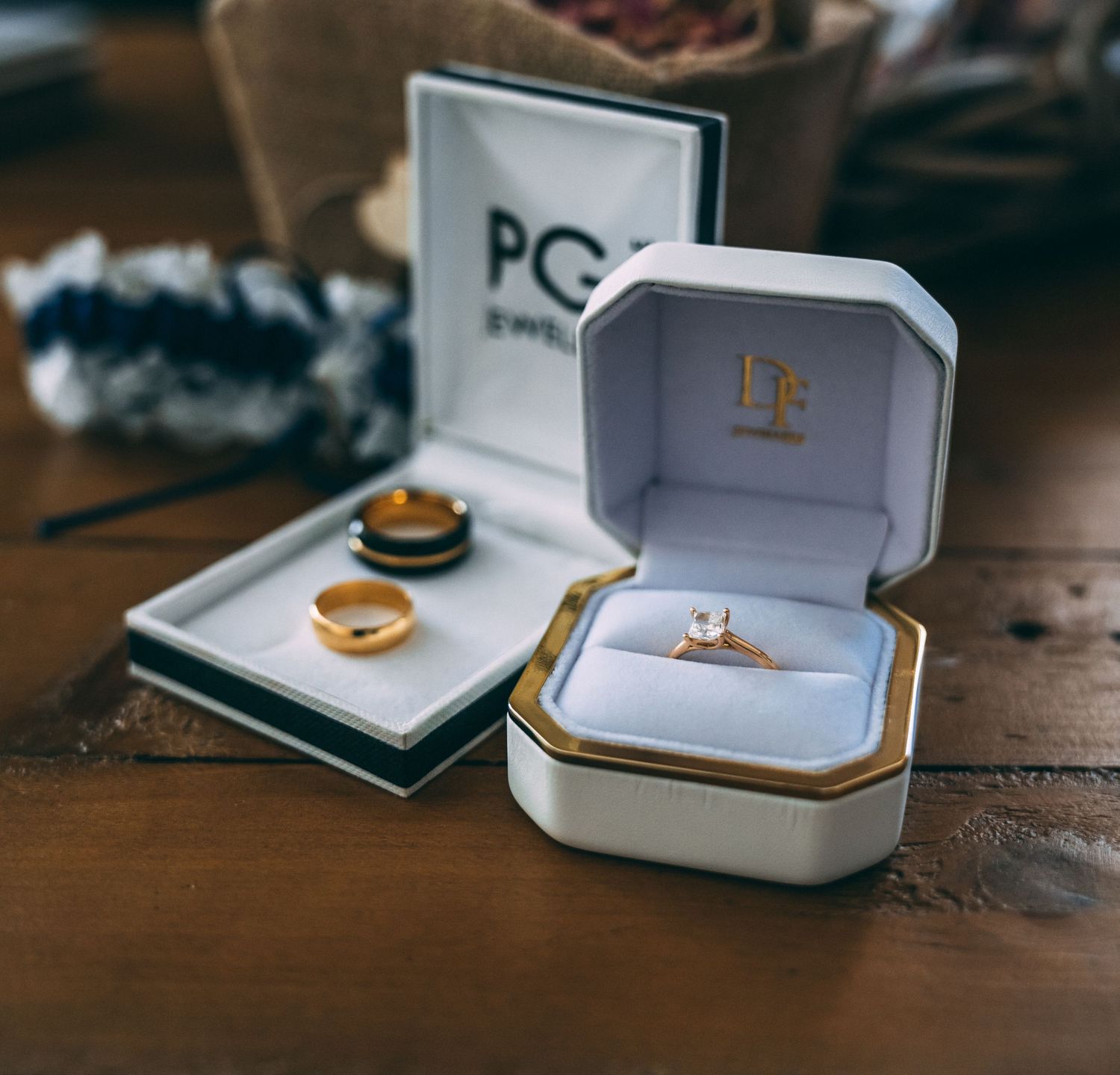 Gold diamond engagement ring in a white luxury jewelry box beside a wedding band on a wooden table.