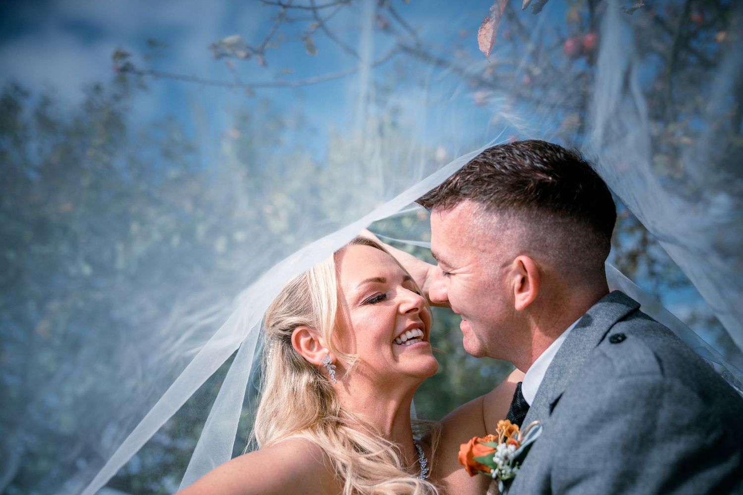 Romantic bride and groom sharing an intimate moment under a flowing veil with a dreamy blue sky backdrop.