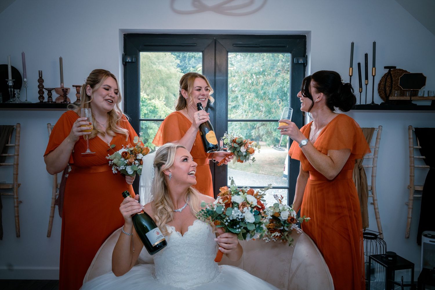 Bride and three bridesmaids in orange dresses celebrating with champagne bottles at a wedding reception.