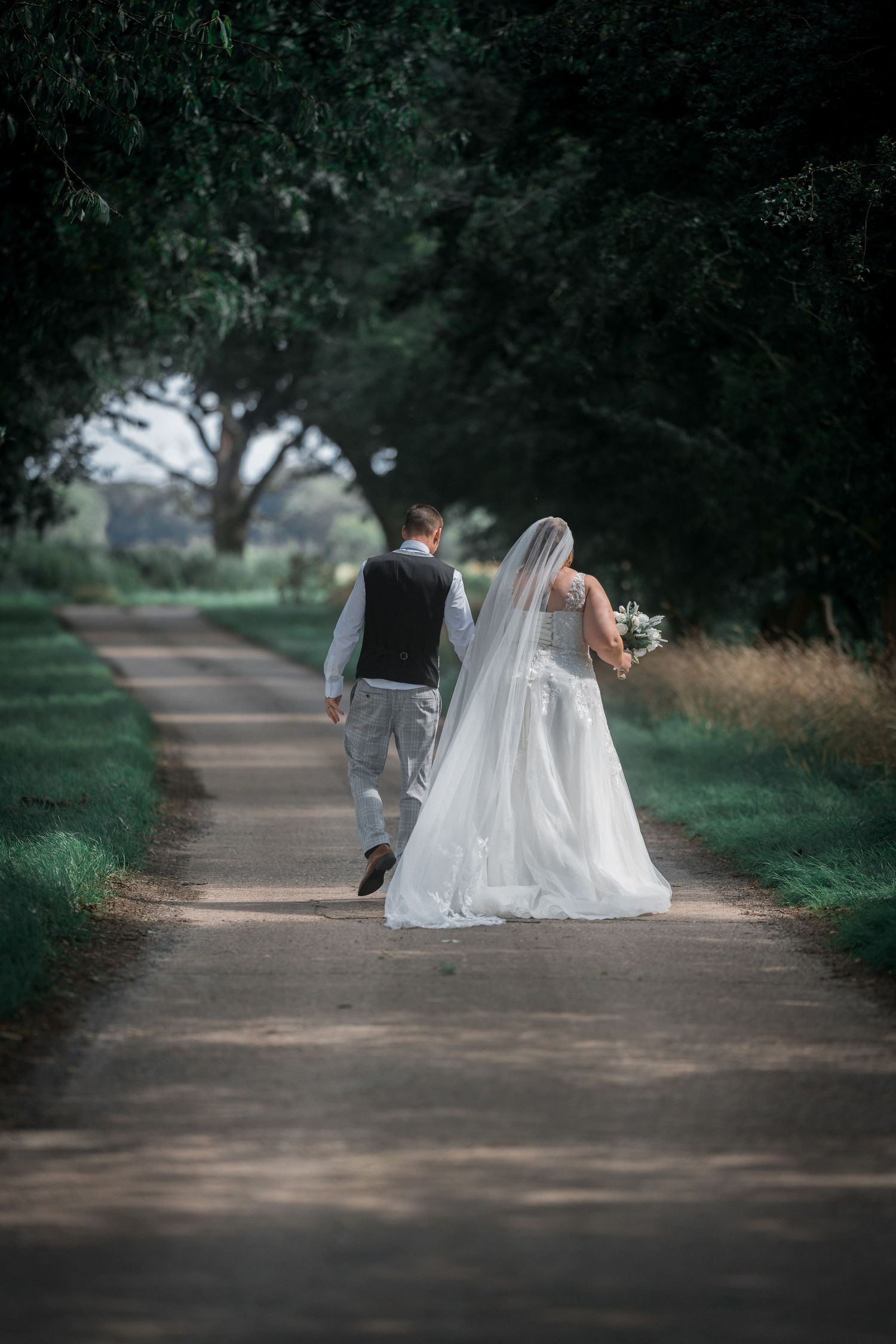 Bride and groom walking hand in hand down a tree-lined path, bride wearing a flowing white gown and veil.