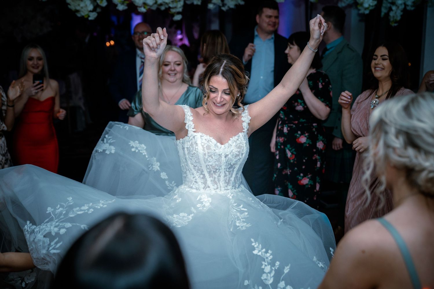 Bride in lace ballgown dancing joyfully with arms raised among guests at wedding reception.