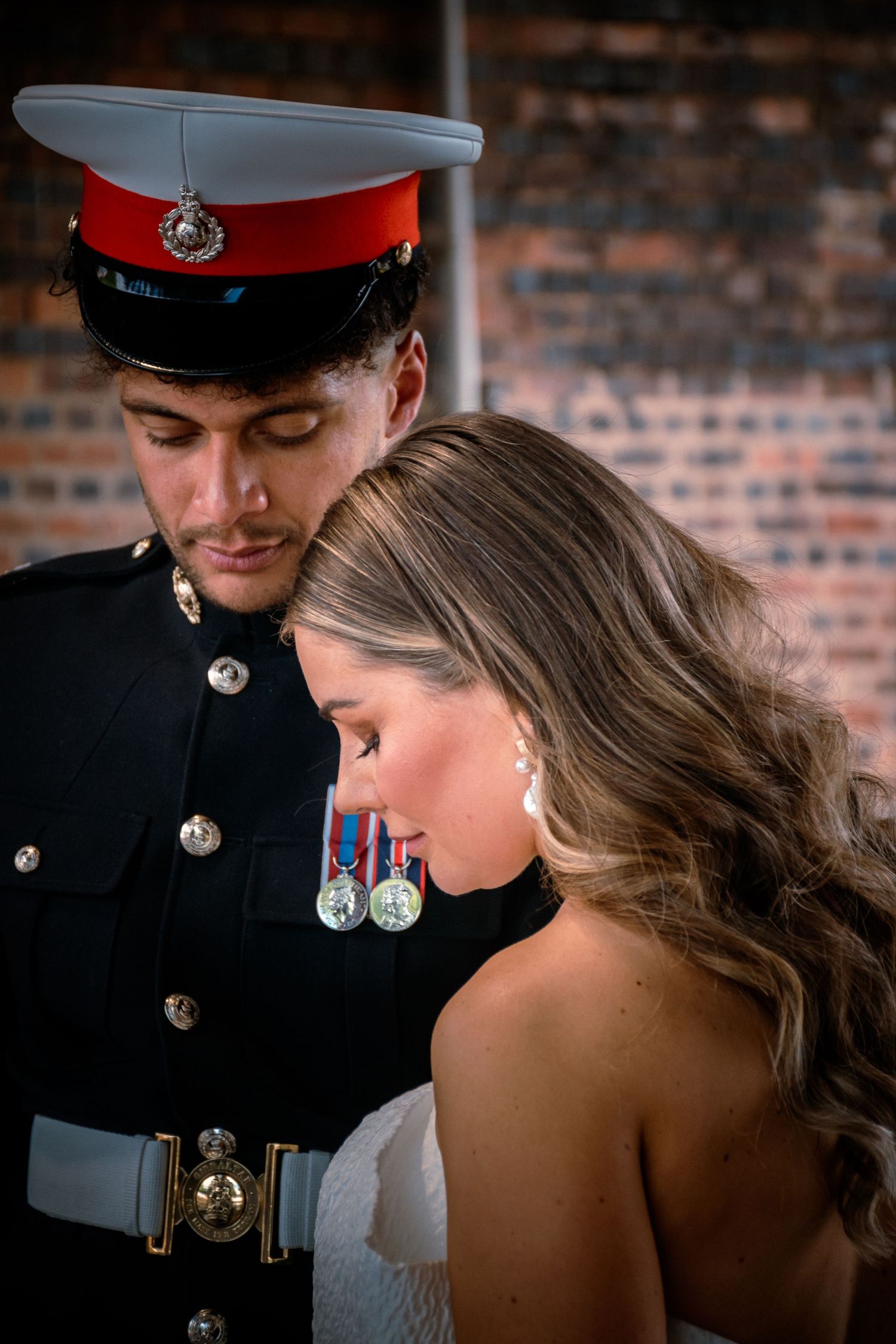 Military groom in dress uniform tenderly looks down at bride against brick wall.