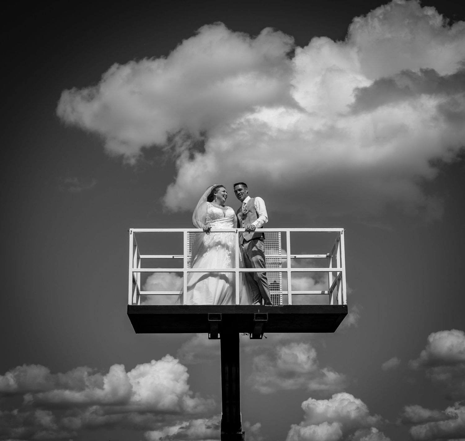 Bride and groom posing on a billboard platform against a dramatic cloudy sky in a black and white wedding photo.