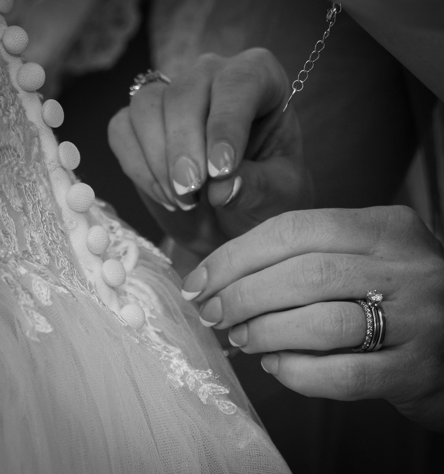 Close-up black and white photo of hands fastening delicate pearl buttons on a lace wedding dress.