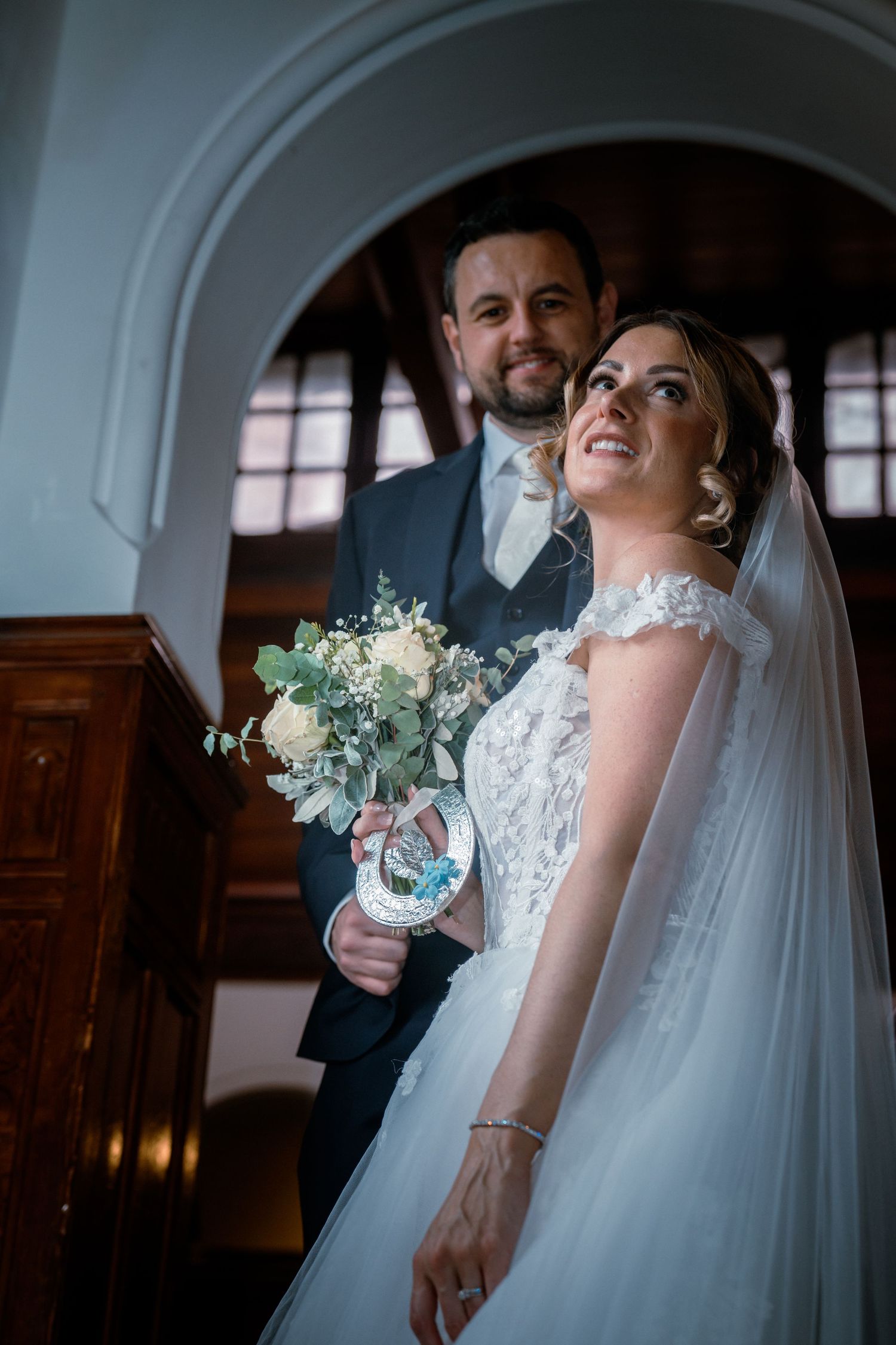 Smiling bride in lace gown holding a white bouquet stands with her groom inside a historic church.