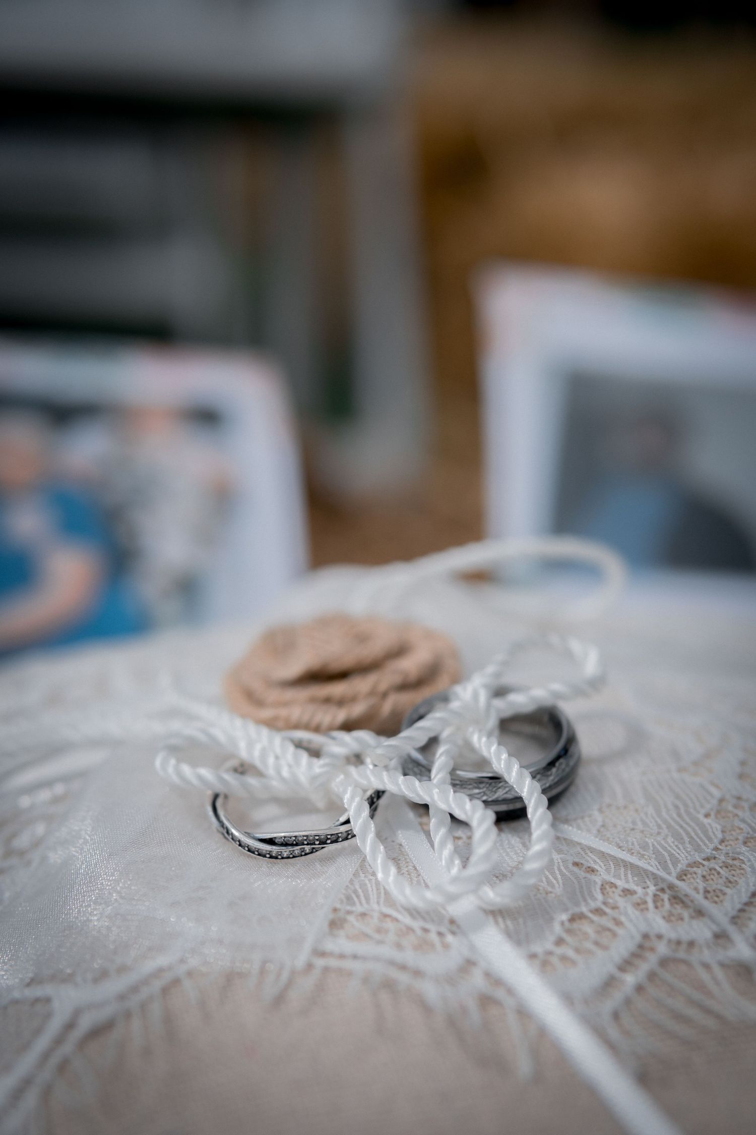 Wedding rings tied with rope on lace pillow with photos in soft background blur.