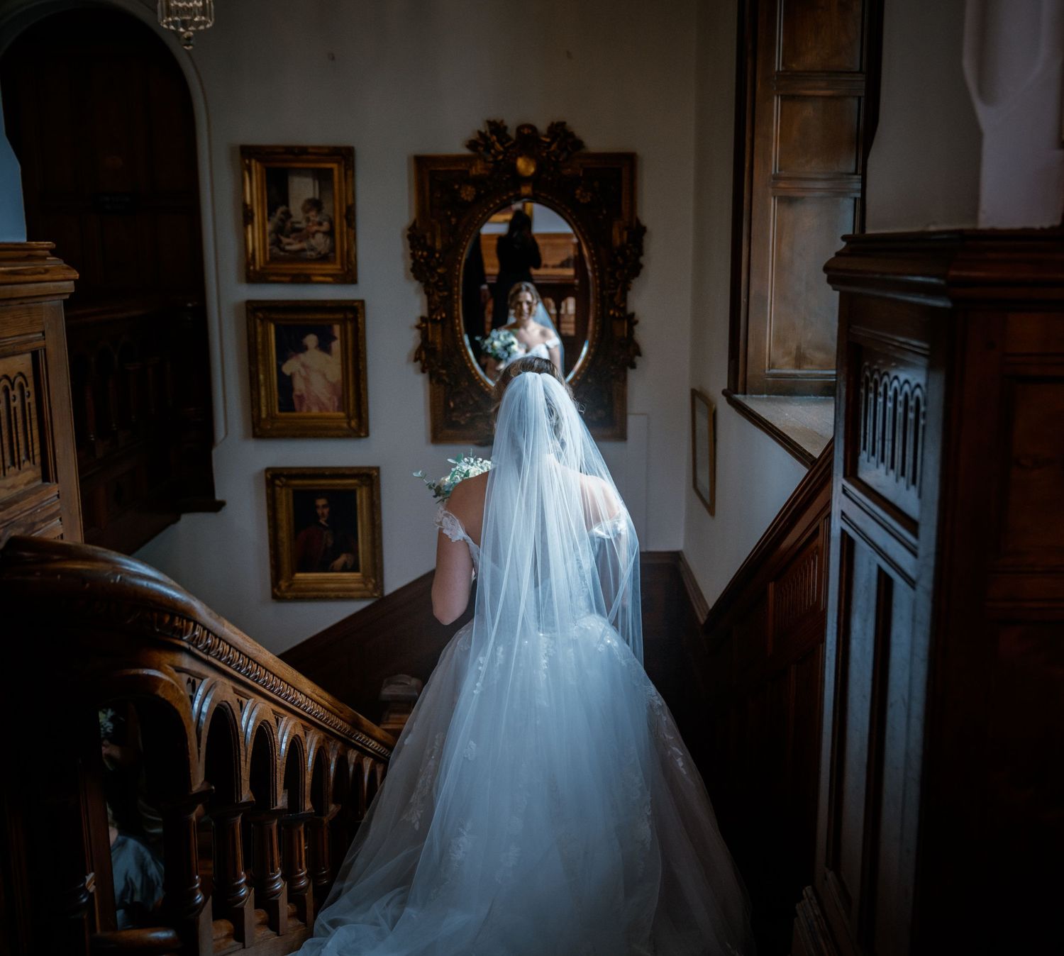 Bride in flowing tulle gown with veil descends grand wooden staircase past antique portraits and ornate mirror.