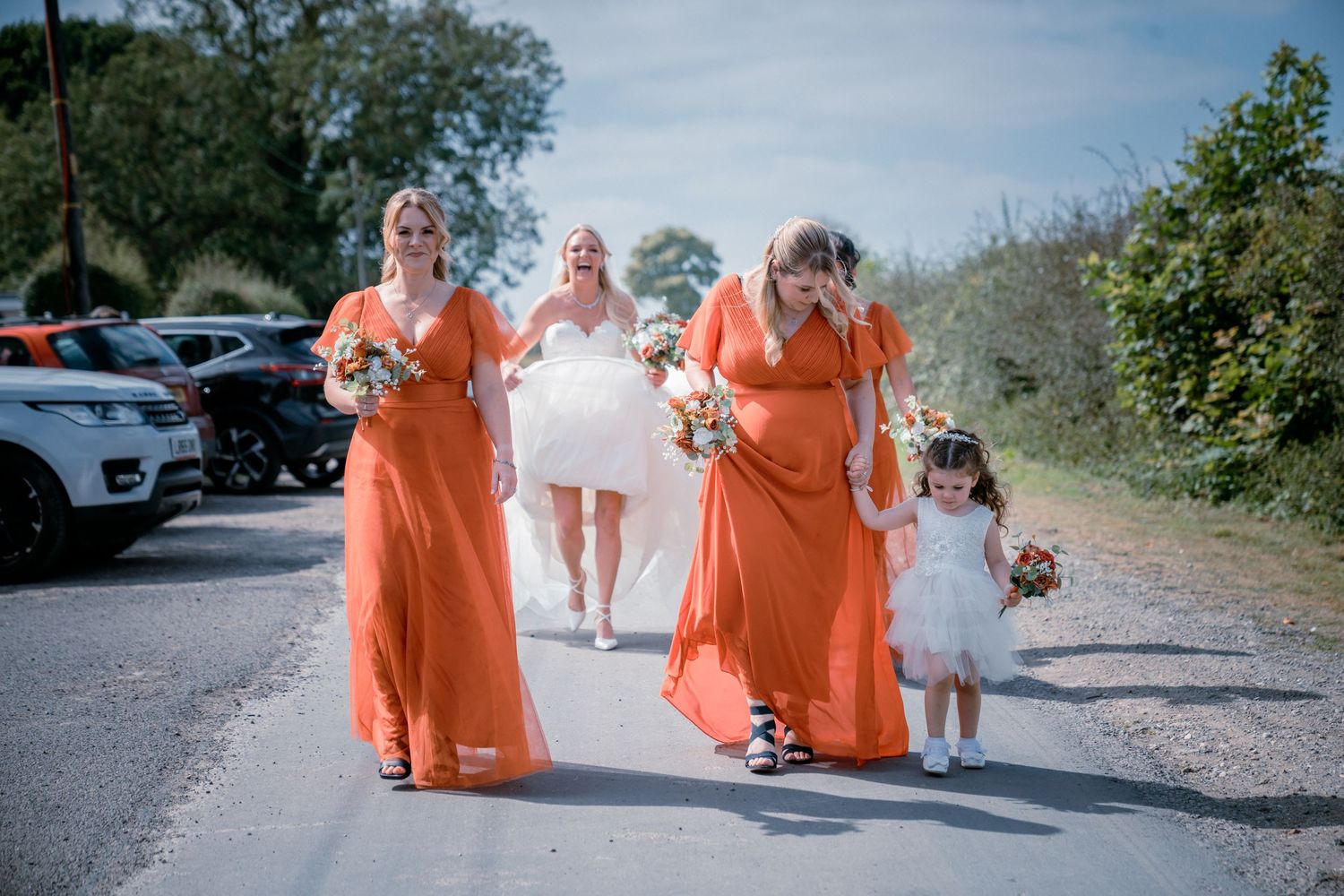 Bridesmaids in orange dresses and flower girl escort bride along sunny outdoor pathway.