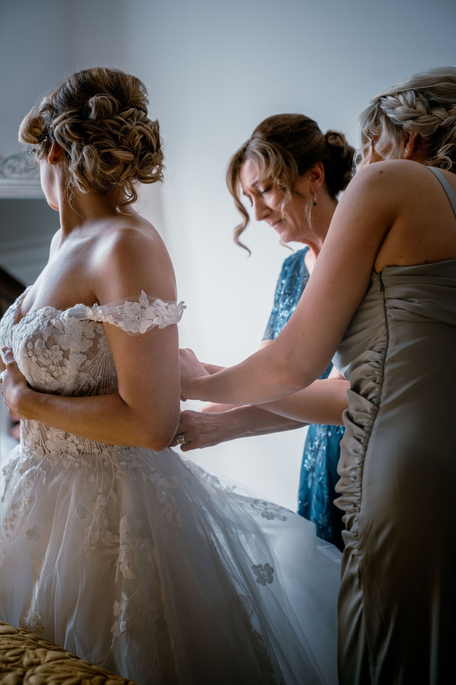 Bridesmaids helping bride fasten her off-shoulder lace wedding gown during bridal preparations.