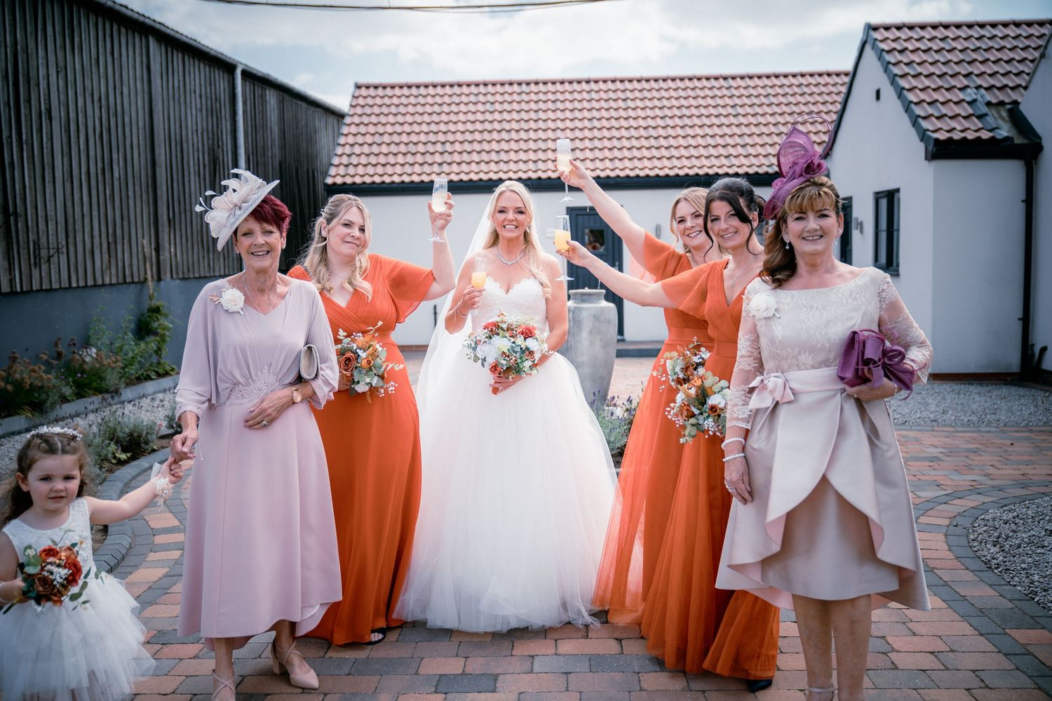 Bride in white ballgown poses with bridesmaids in orange and pink dresses outside rustic barn venue.