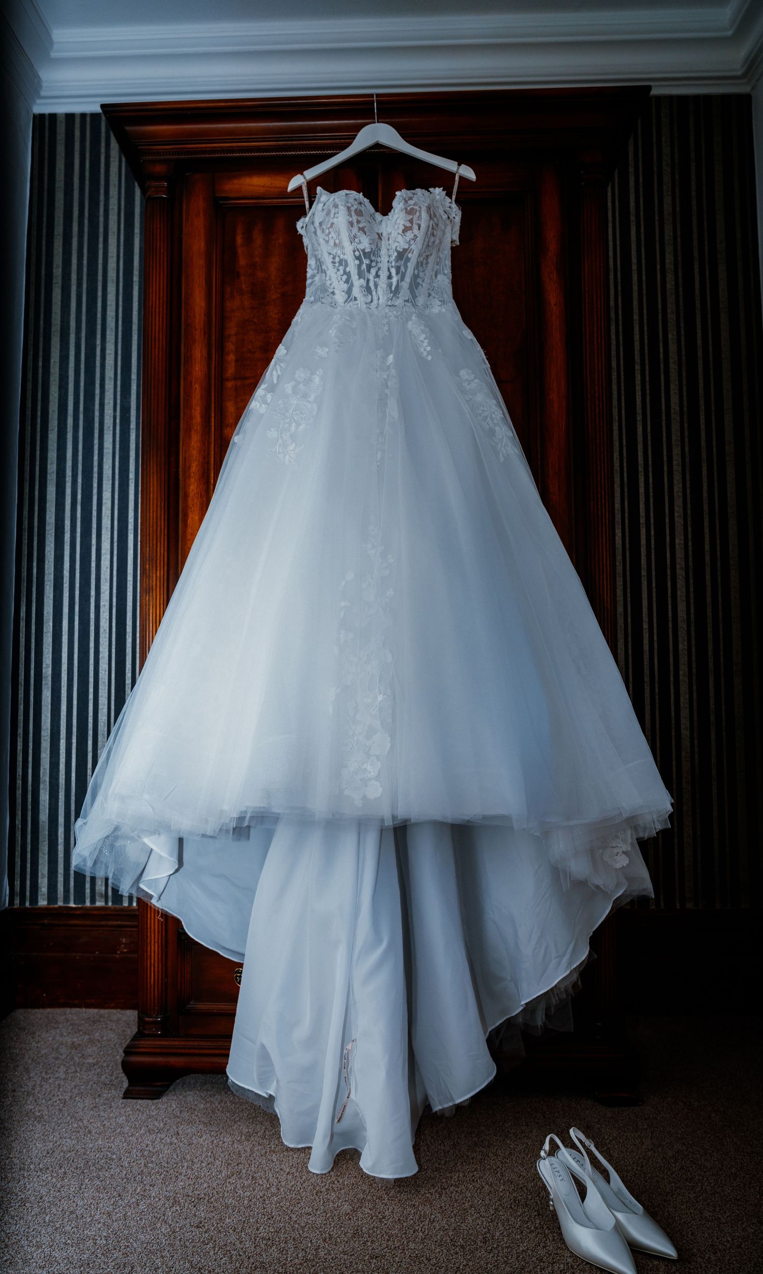 Elegant strapless white lace ballgown wedding dress hanging on a wooden wardrobe with bridal heels below.