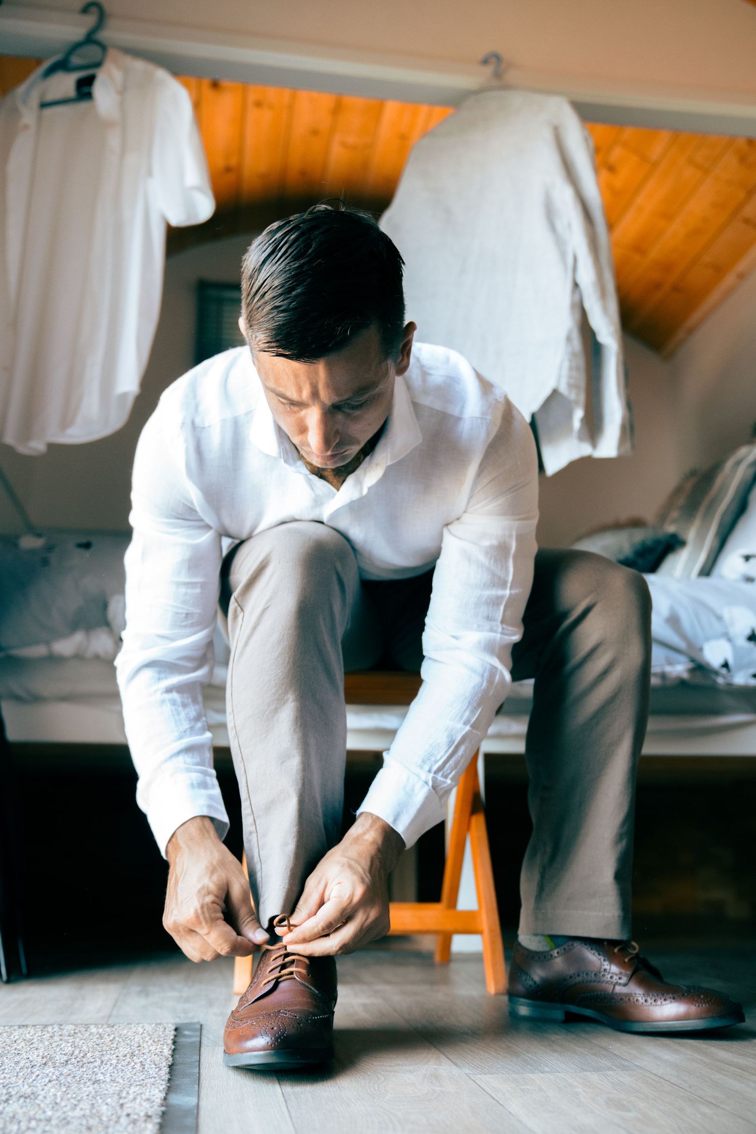 Groom in white shirt and grey trousers tying his brown leather shoes while seated indoors before wedding.