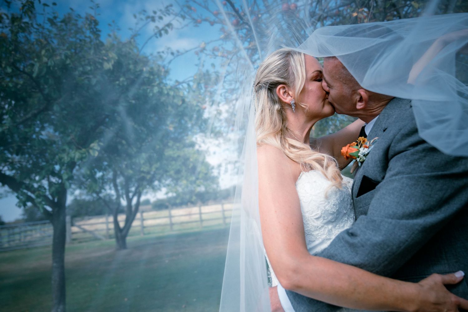 Bride and groom sharing romantic kiss under flowing veil outdoors on wedding day.