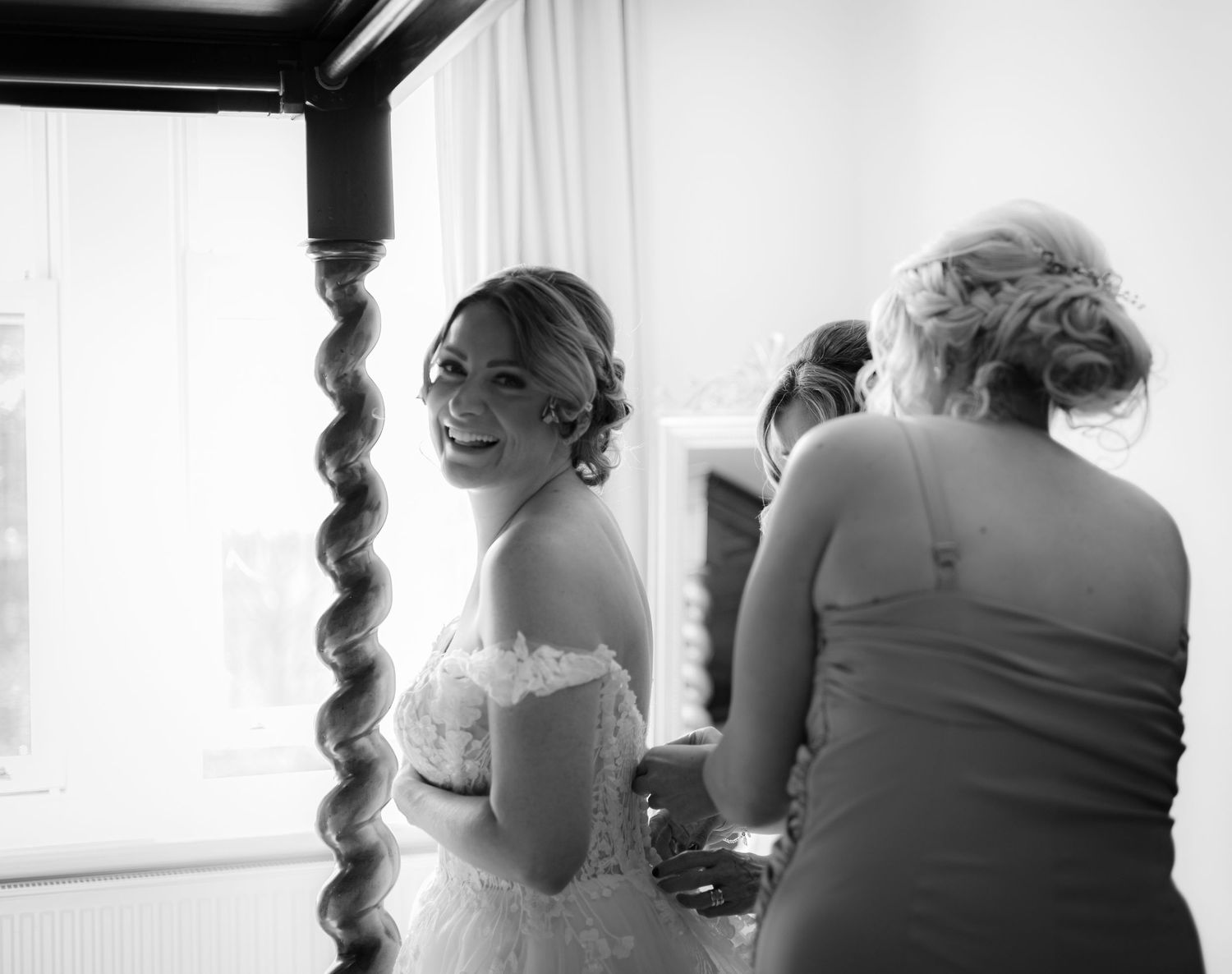 Black and white photo of laughing bride in lace gown being helped by attendants during preparations.