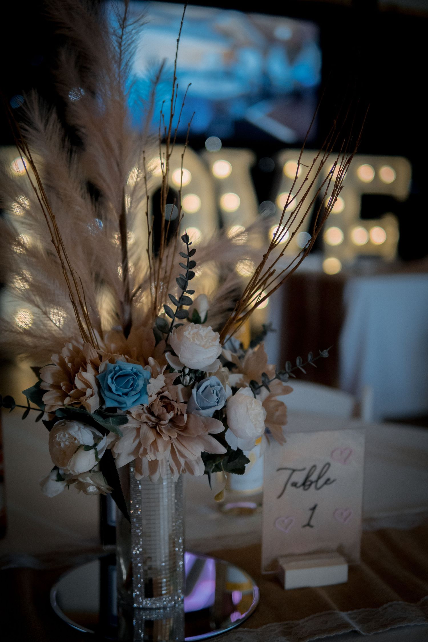Wedding table centerpiece with pampas grass, blue and white roses beside a lit LOVE sign.