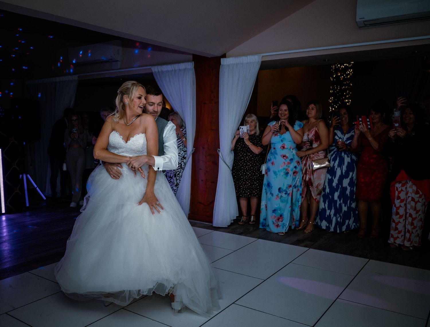 Bride and groom sharing their first dance on a white dance floor surrounded by cheering wedding guests.
