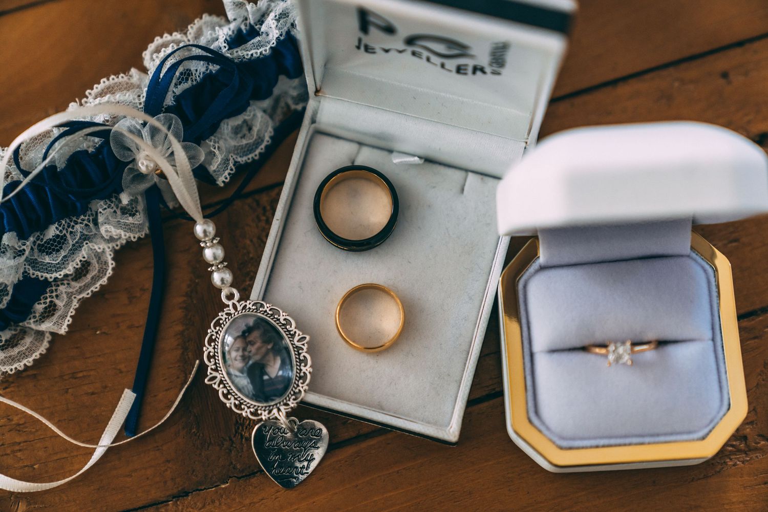 Wedding rings, diamond engagement ring, garter, and heart locket displayed on wooden table.