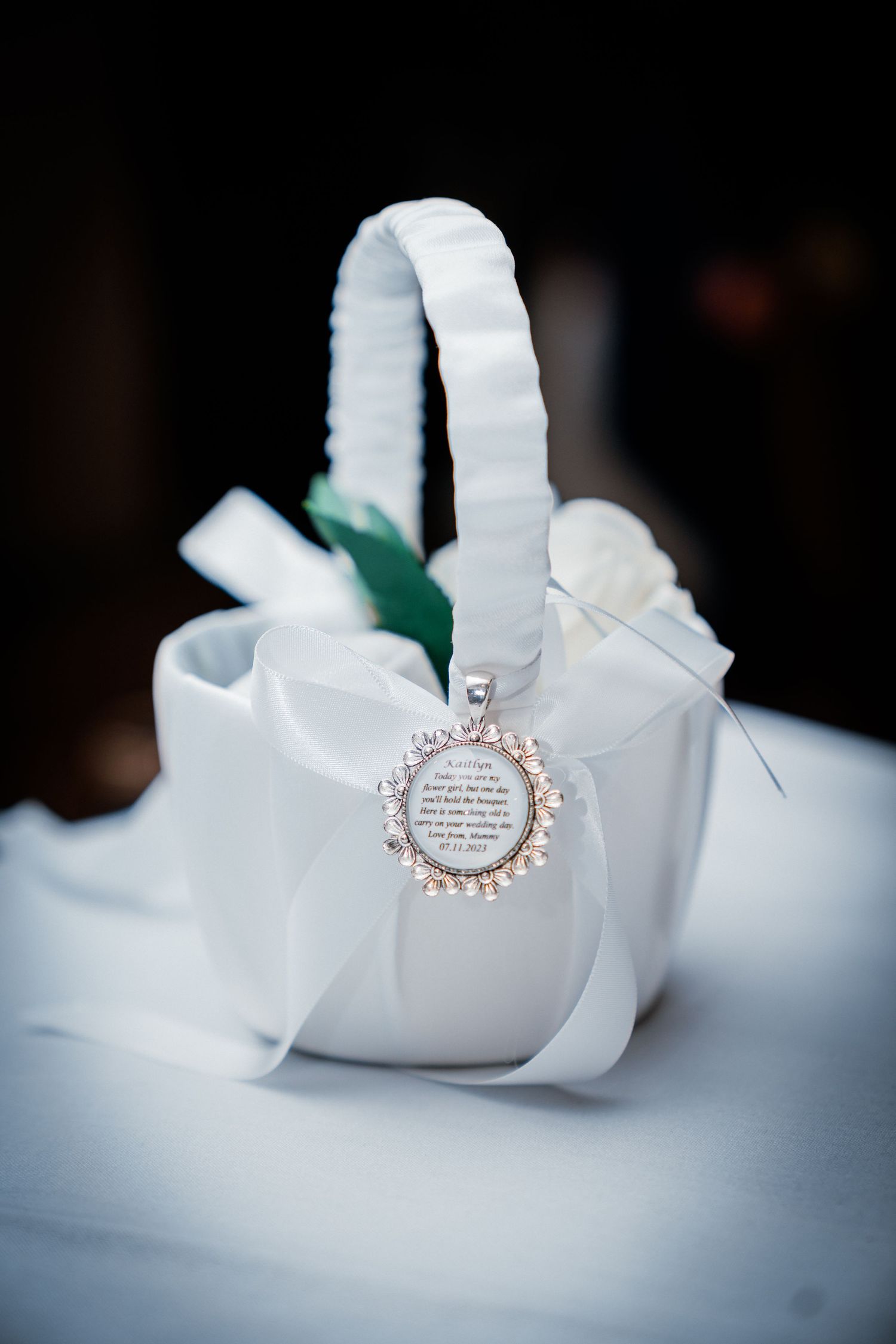 White satin flower girl basket with a personalized circular charm and ribbon bow on a white surface.