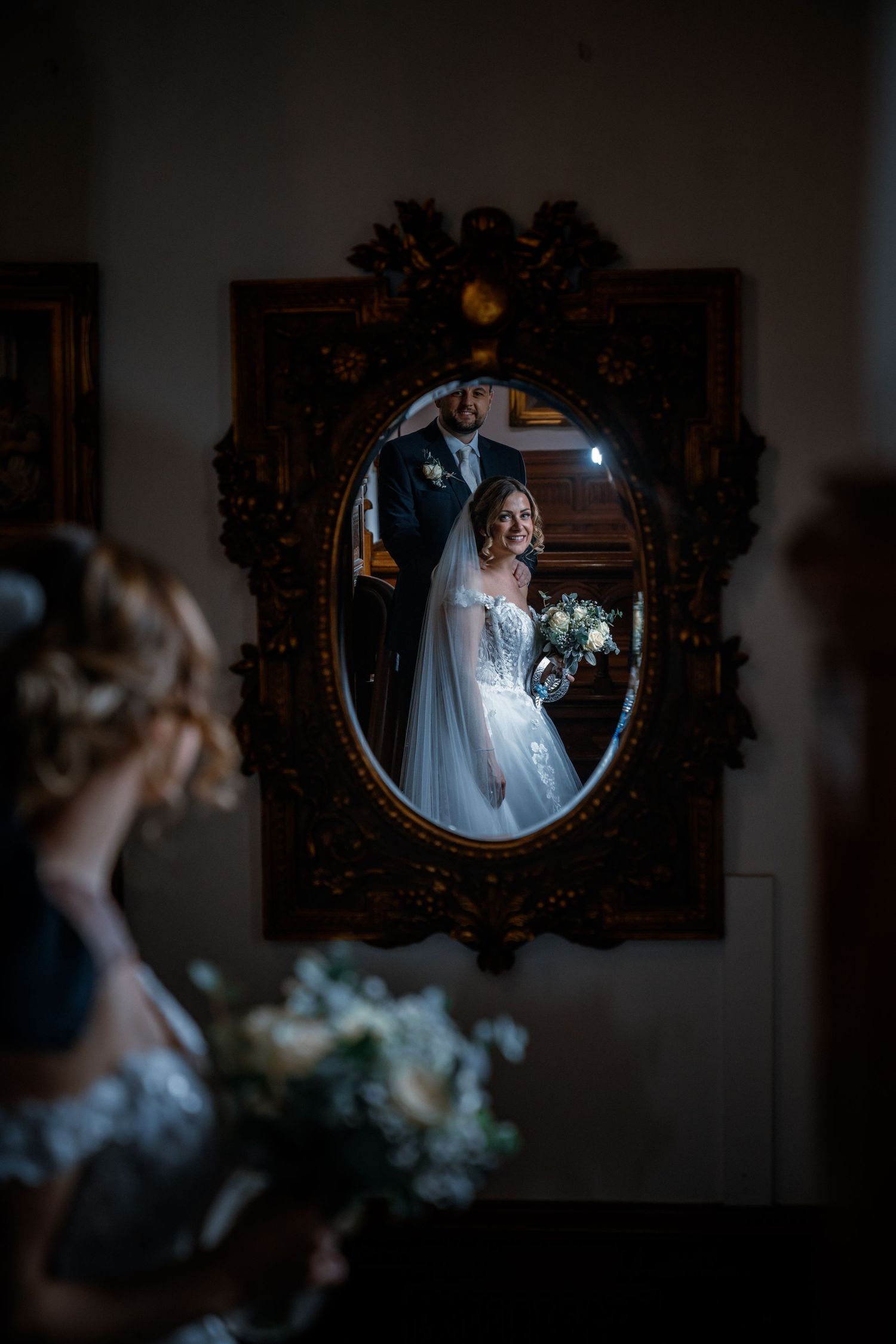 Bride and groom reflected in ornate vintage mirror, bride holding lush white bouquet on wedding day.