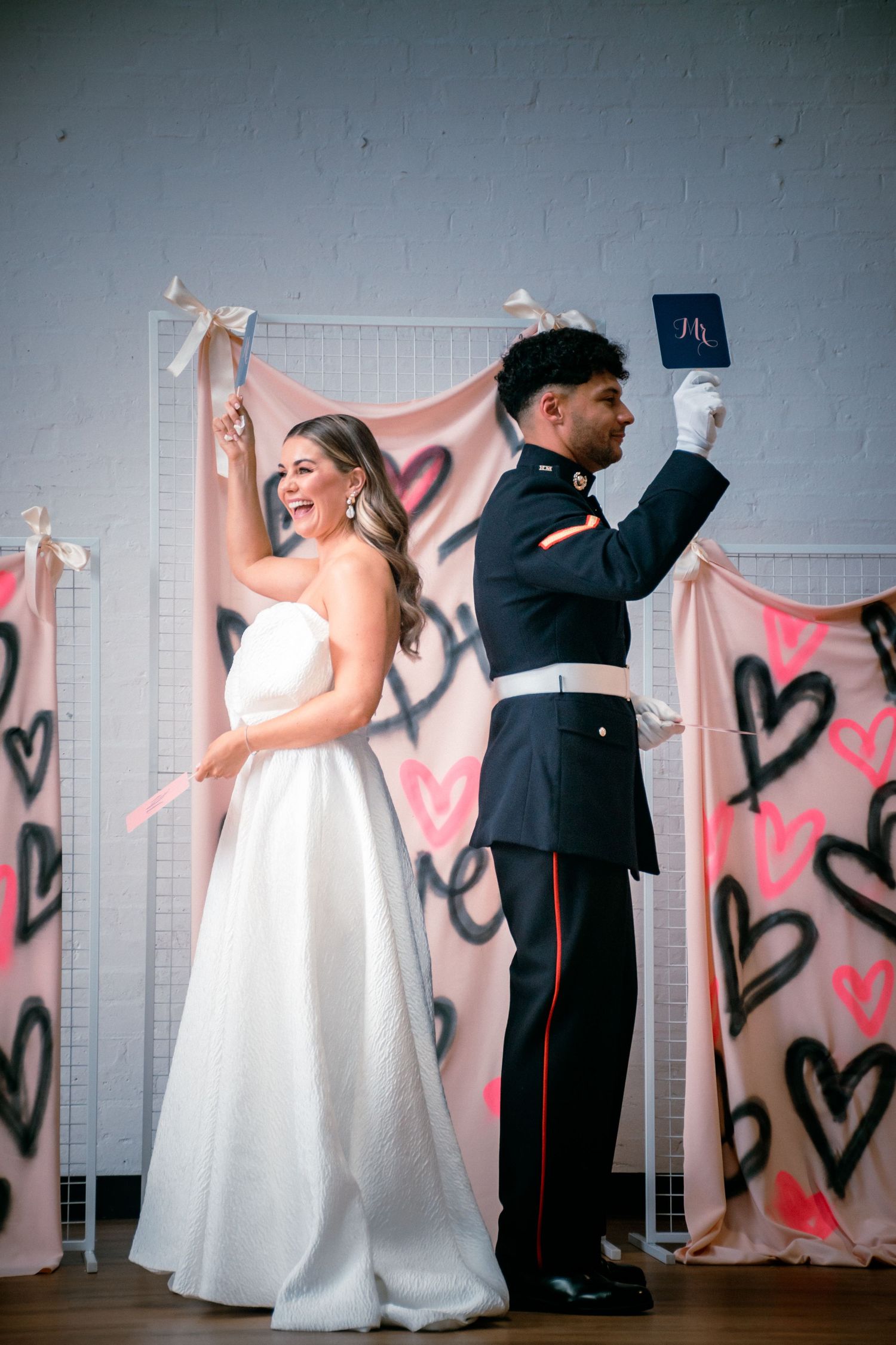 Bride and military groom stand back-to-back holding vow books before heart backdrop.