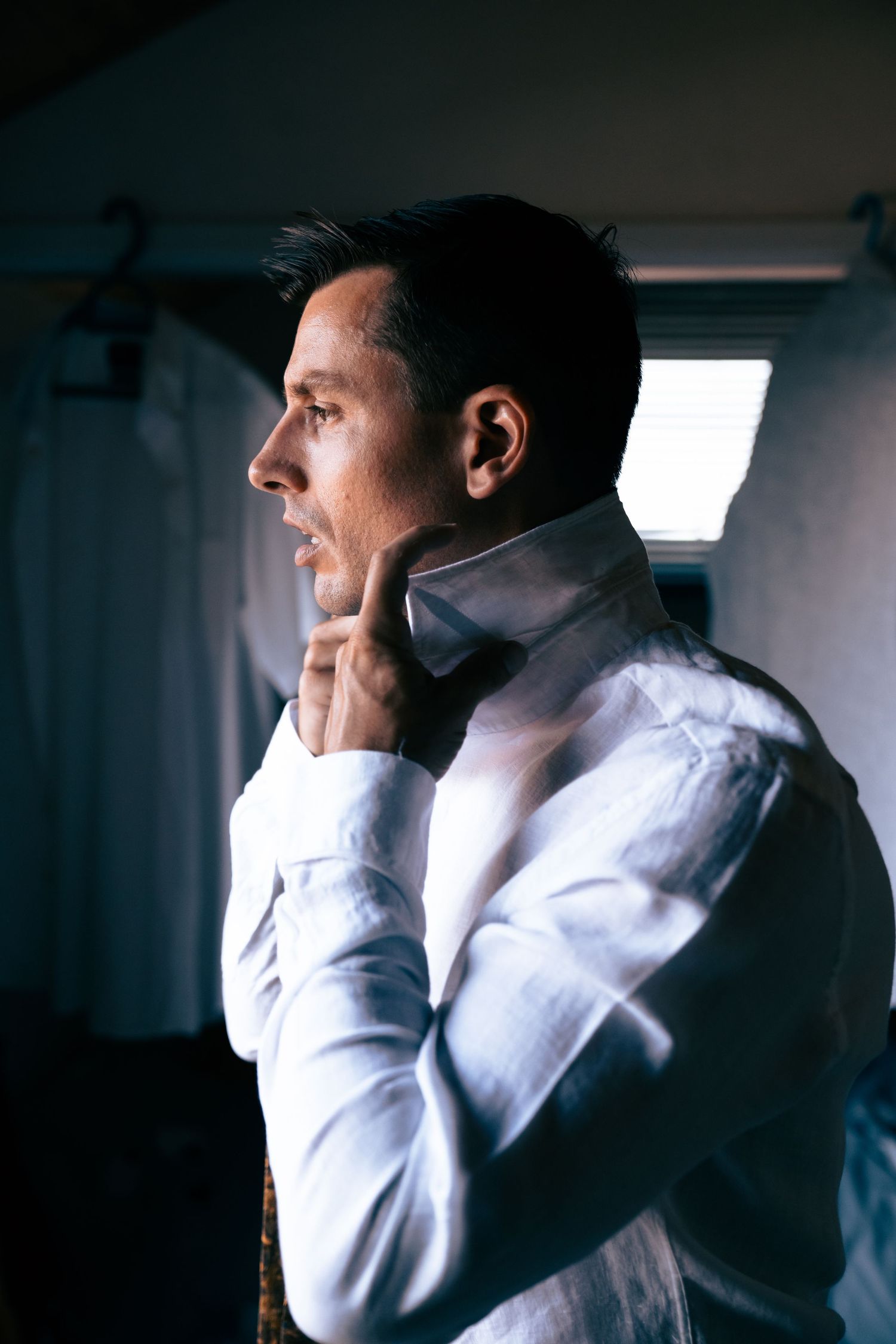 Groom in white dress shirt adjusting his collar while looking out a window before his wedding ceremony.