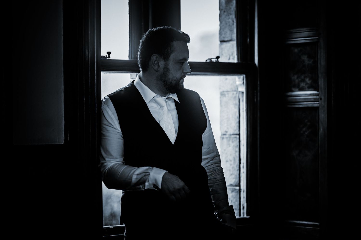 Groom in white shirt and black vest standing by window in moody black and white wedding photo.