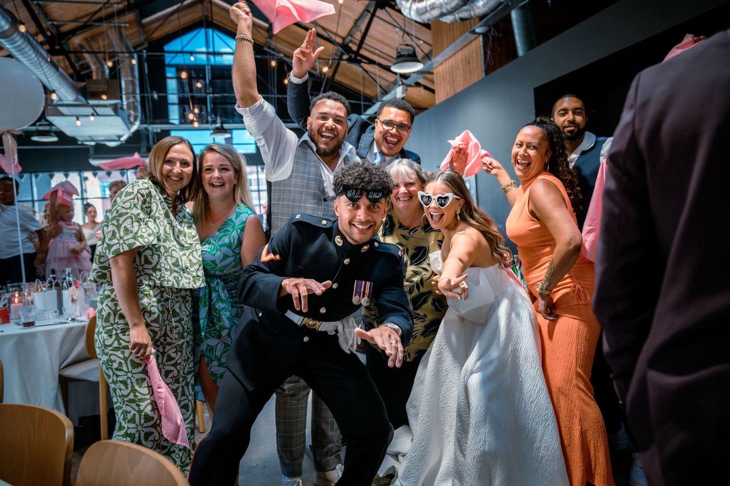 Joyful wedding party group photo with bride and groom surrounded by celebrating guests at an indoor reception venue.
