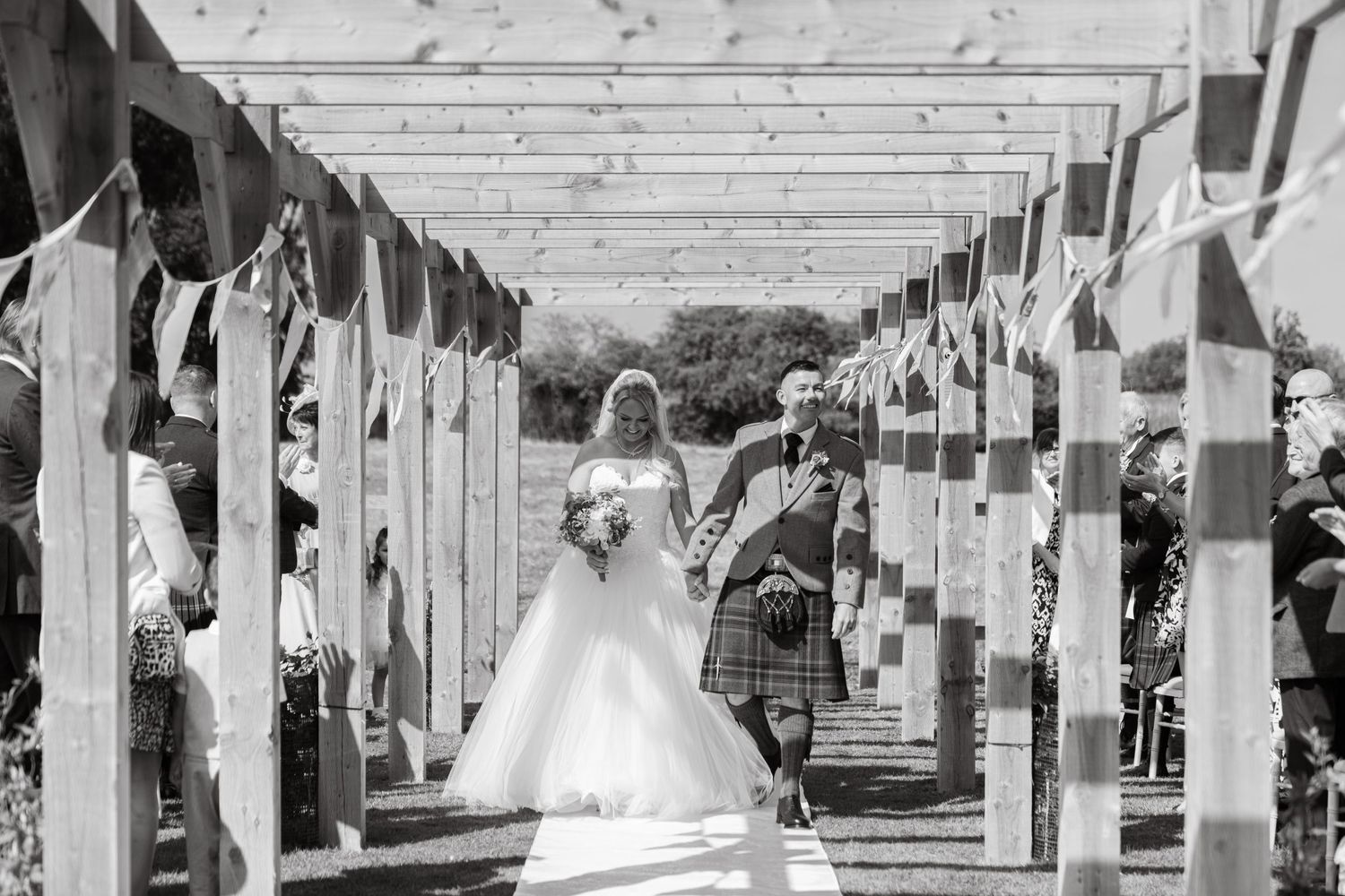 Bride walks down outdoor aisle with father in kilt, flanked by wooden pergola and bunting, black and white wedding photo.