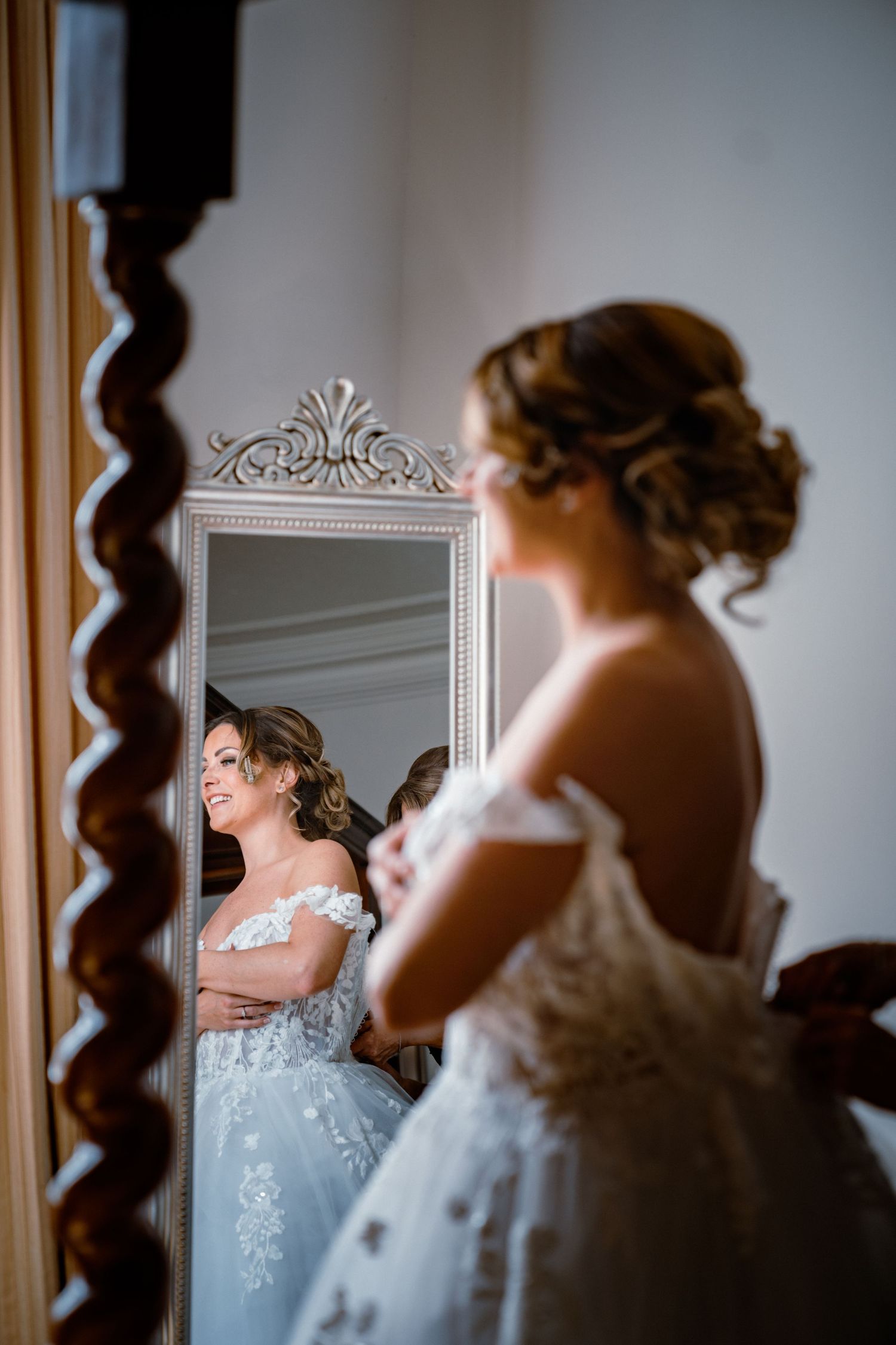 Bride in off-shoulder lace gown smiles at her reflection in an ornate white-framed full-length mirror.