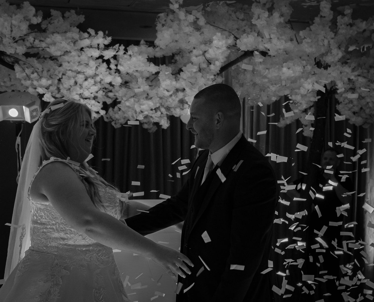 Black and white photo of bride and groom sharing first dance under floral arch with confetti falling.