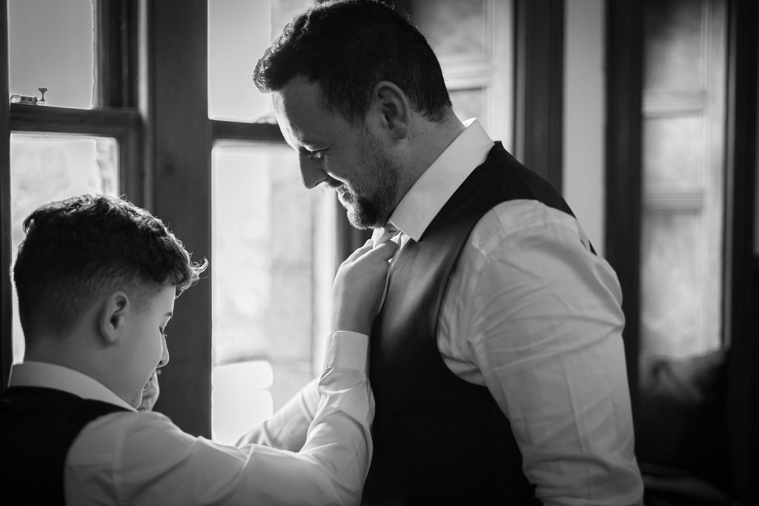 Groom in black vest adjusting bow tie with help from young boy in black and white photo.