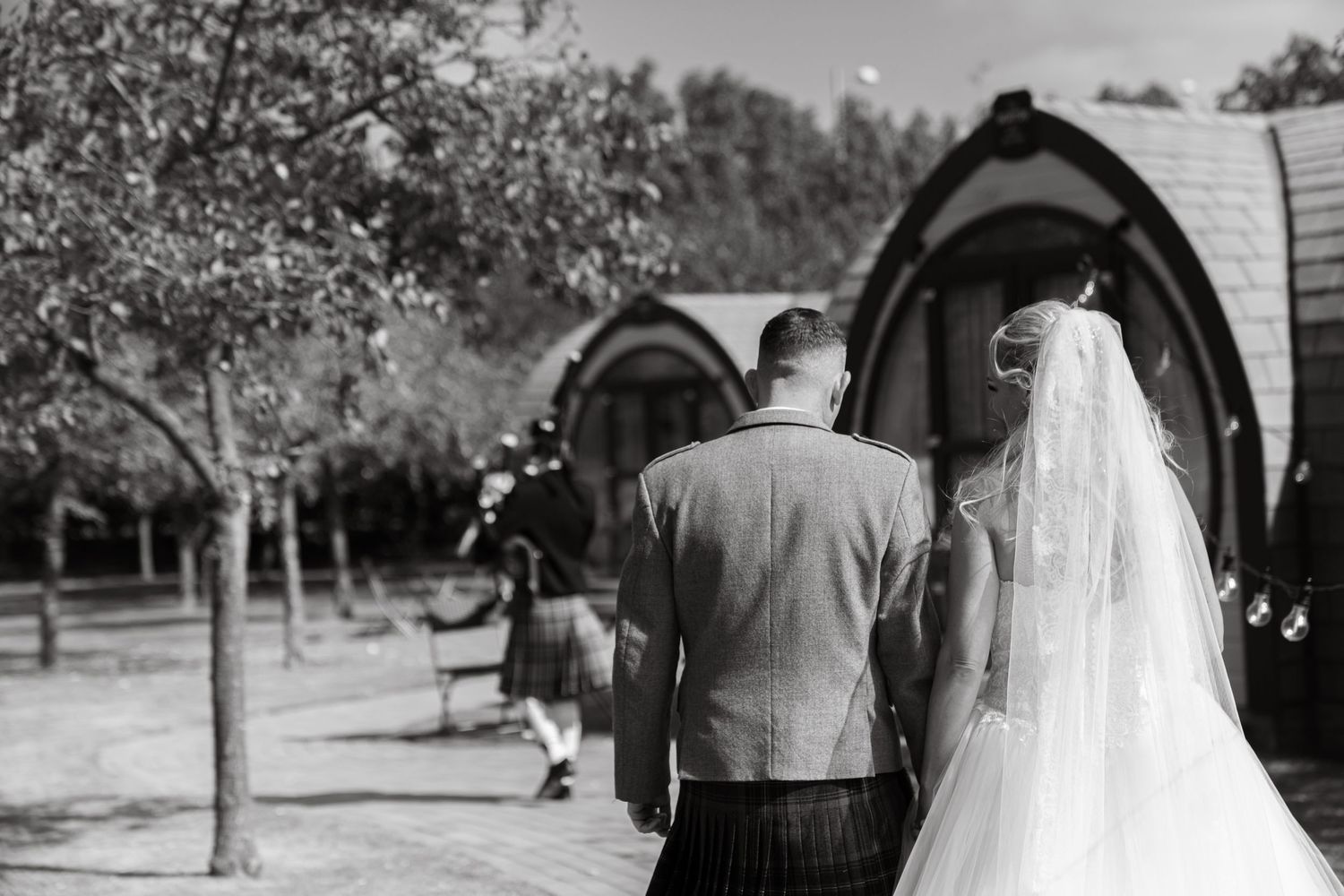 Black and white rear view of bride and groom in kilt walking toward rustic barrel wedding venue.