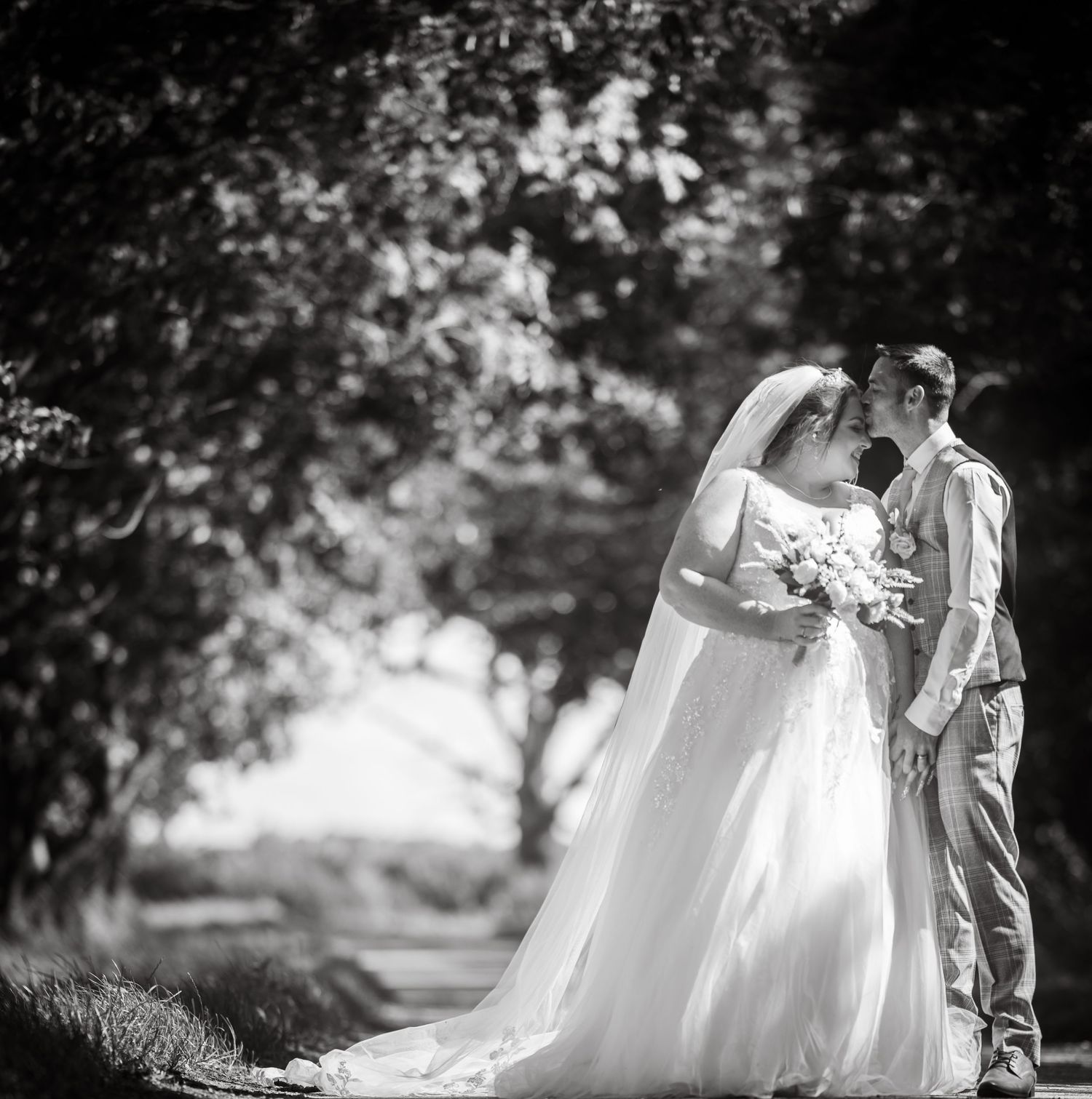 Black and white portrait of bride and groom standing close together under lush trees with bouquet.