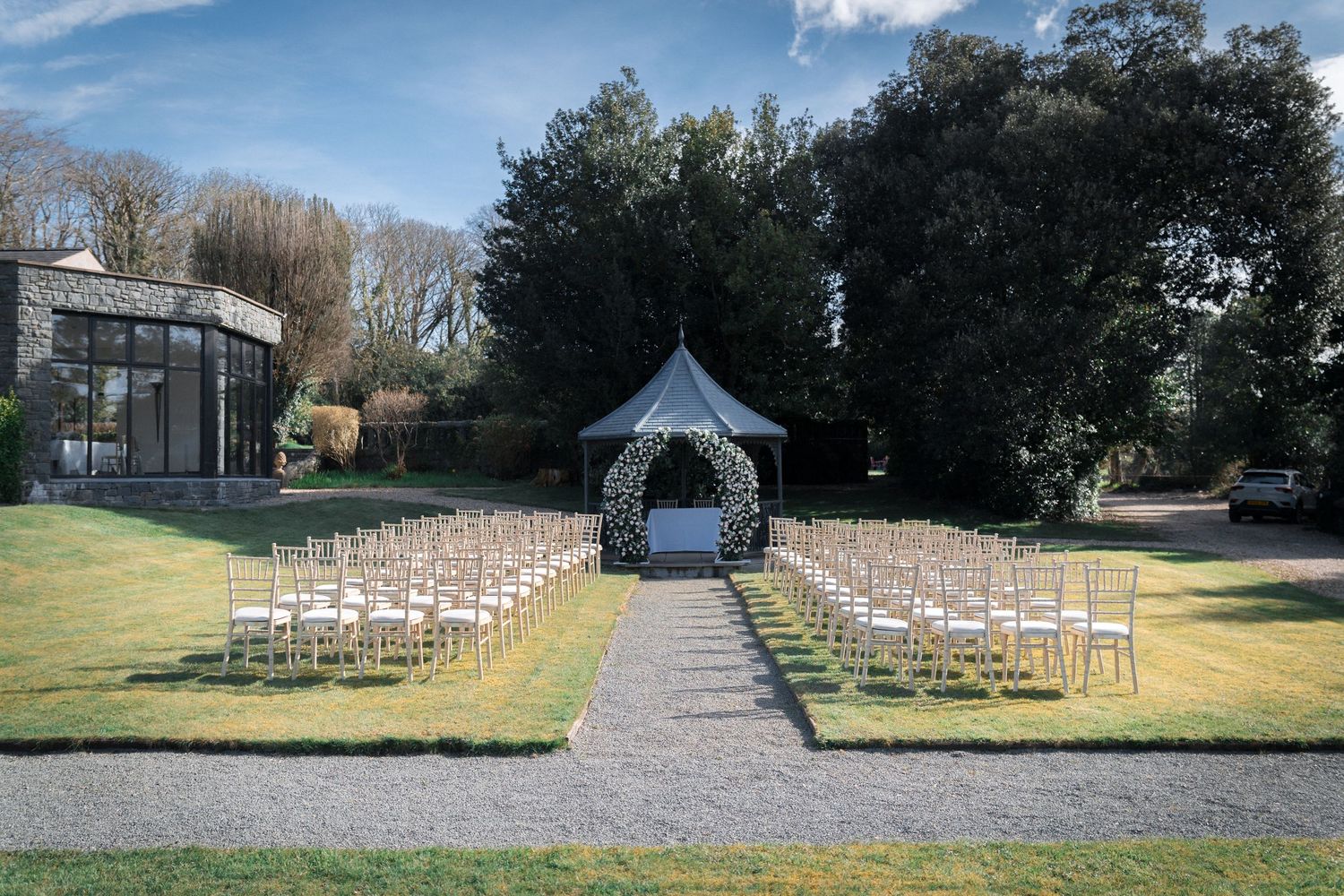 Outdoor wedding ceremony setup with white chairs and floral gazebo arch on manicured lawn.