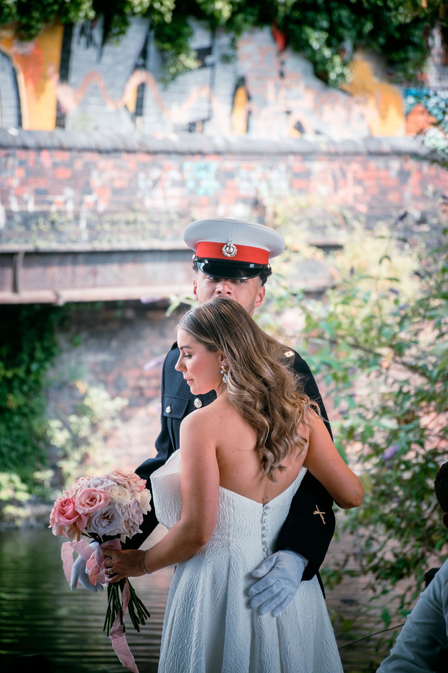 Bride in a strapless gown holding a pink bouquet beside a groom in military uniform near a river.